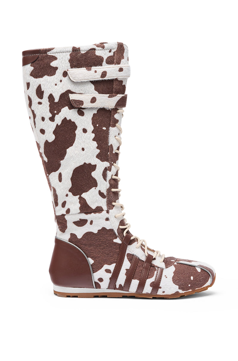 The AZALEA WANG OZZY BROWN MULTI FLAT KNEE HIGH SNEAKER features a brown and white cow print in faux pony hair, white laces, a top velcro strap, brown heel and toe accents, and a flat sole. Shown from the side view. 3