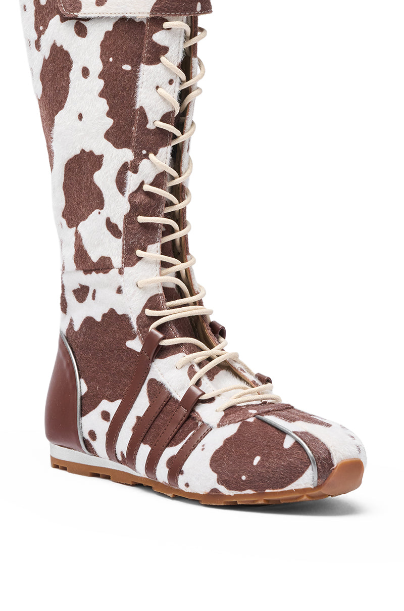 The AZALEA WANG OZZY BROWN MULTI FLAT KNEE HIGH SNEAKER features a brown and white cow print in faux pony hair, leather accents on the sides and heel, and a flat sole for comfort. 7