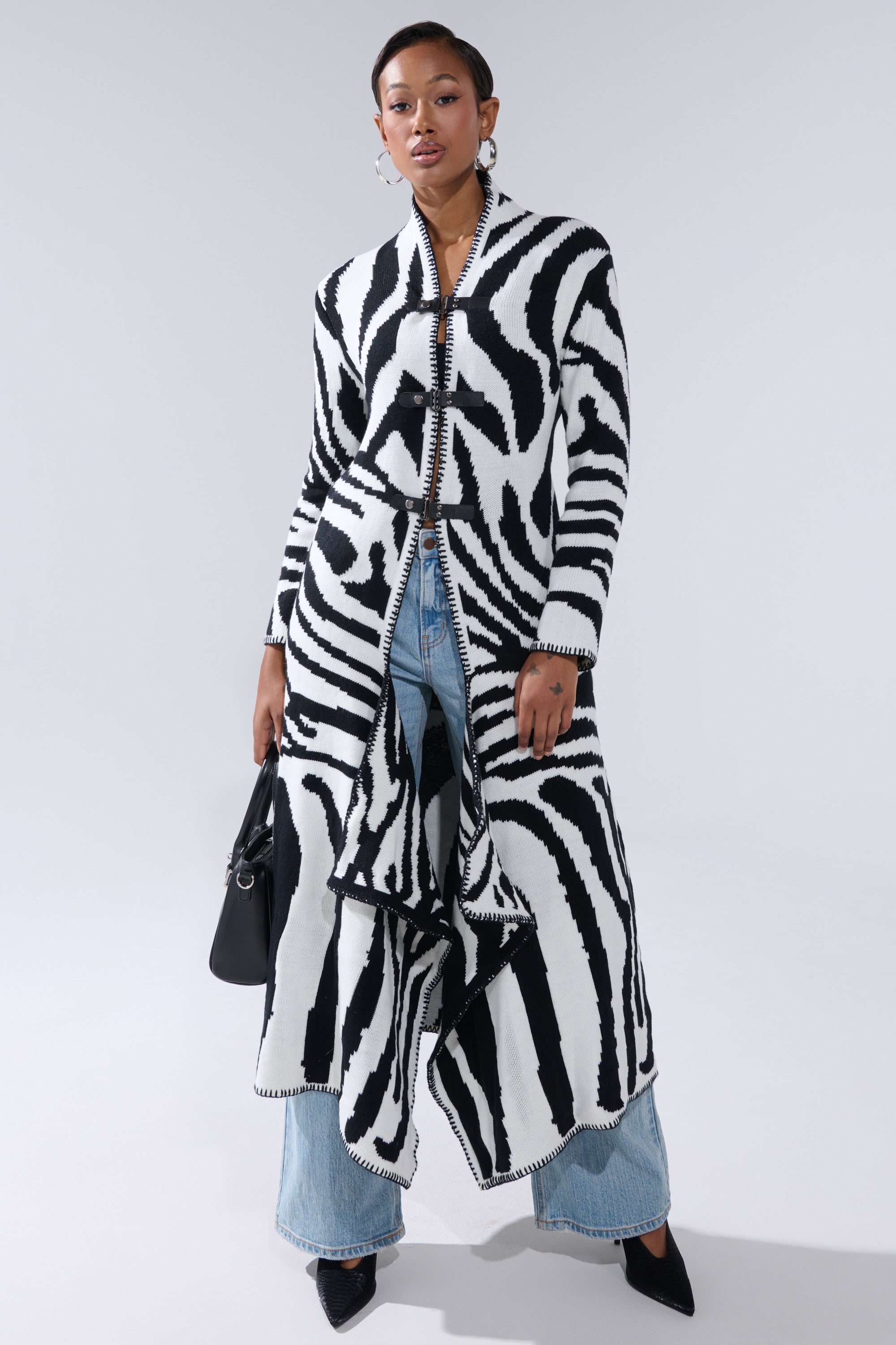 Confidently posing against a gray background, a woman wears THE RICH BITCH IS HERE LONG CARDIGAN—a longline zebra print style with faux leather strap closures—over light blue jeans and black heels, accessorized with a black handbag.
