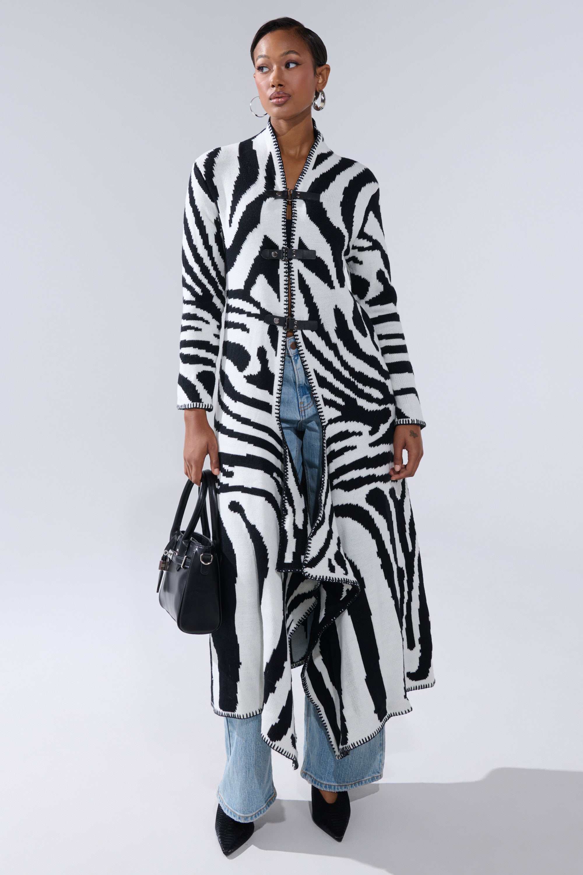 A woman wears THE RICH BITCH IS HERE LONG CARDIGAN with zebra print over light blue jeans and black pointed shoes, holding a black handbag with a faux leather strap against a plain light background.