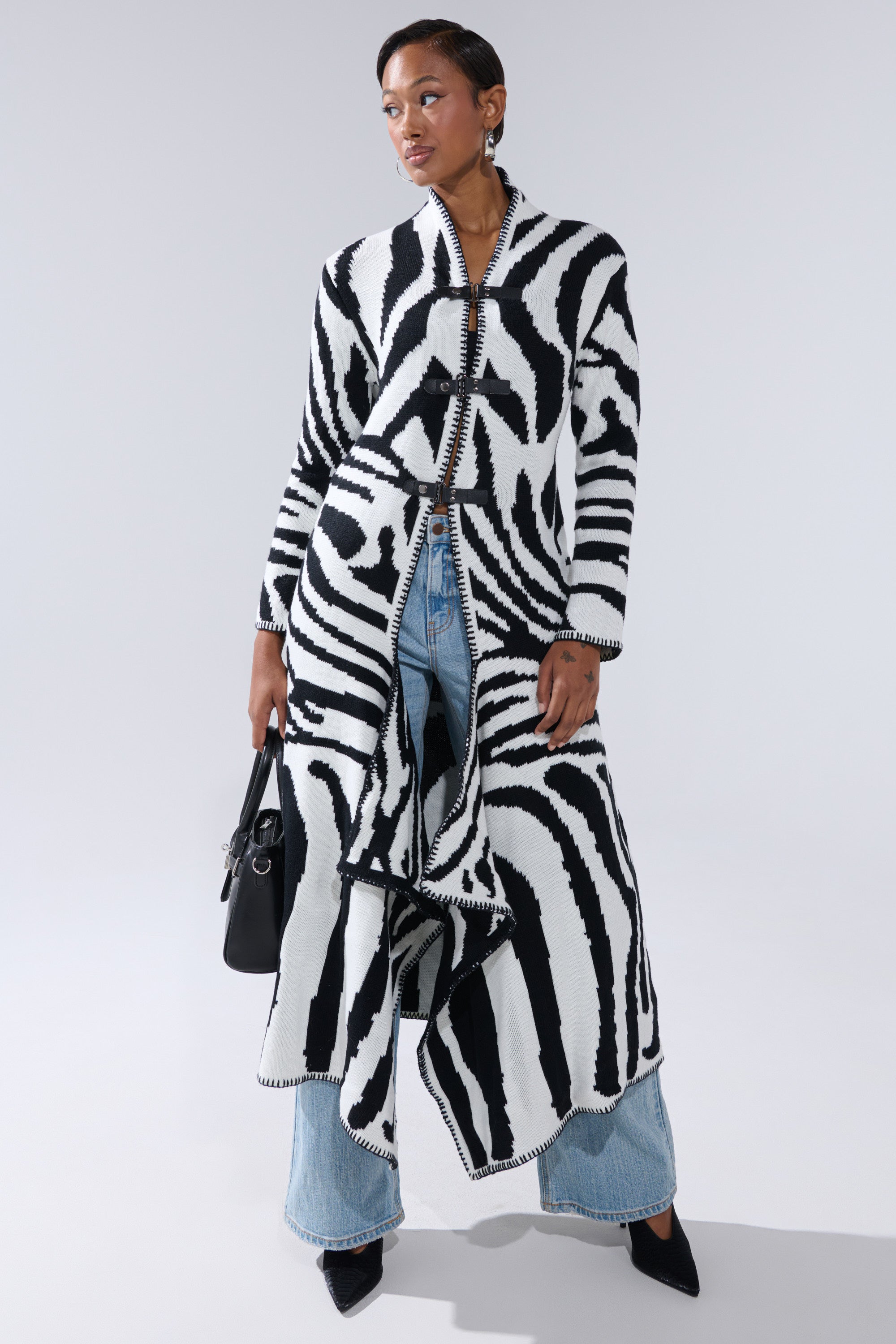 A woman stands against a light gray background wearing THE RICH BITCH IS HERE LONG CARDIGAN over blue jeans and black pointed shoes, holding a black faux leather handbag.