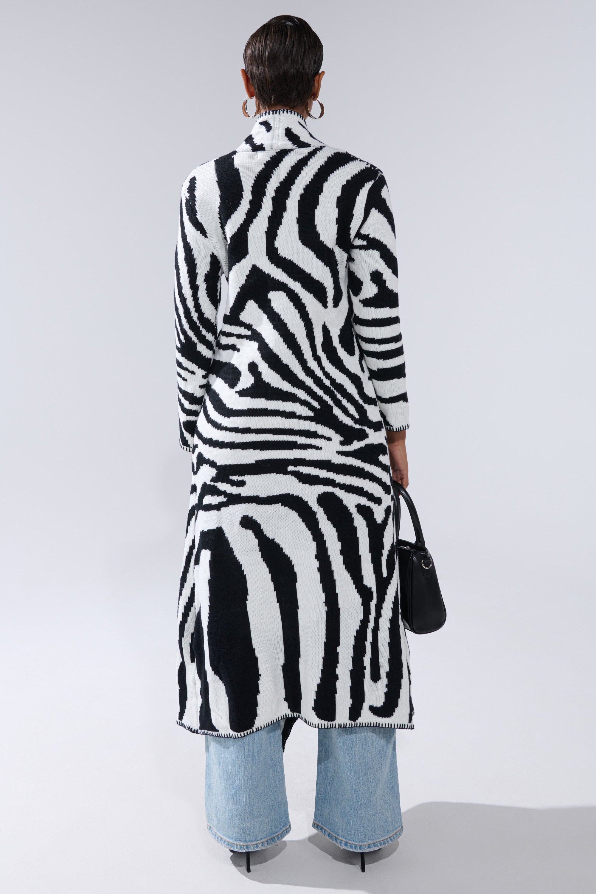 A person stands facing away, wearing THE RICH BITCH IS HERE LONG CARDIGAN with a zebra print over light blue wide-leg jeans, holding a black handbag with a faux leather strap against a plain light background.