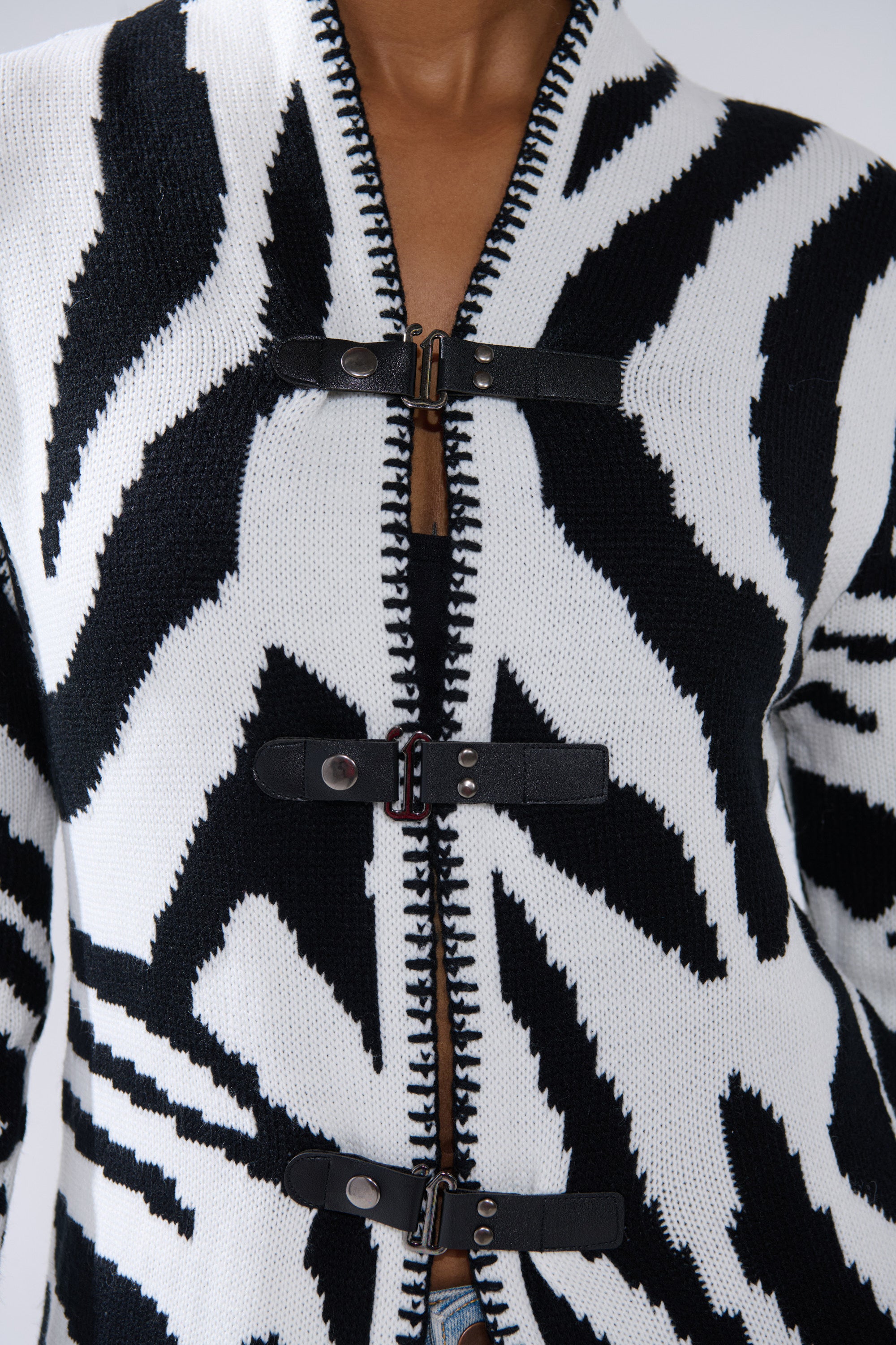 Close-up of a person wearing THE RICH BITCH IS HERE LONG CARDIGAN, a bold zebra print jacket with three faux leather strap closures over a zipper. This graphic black and white piece features an eye-catching design.