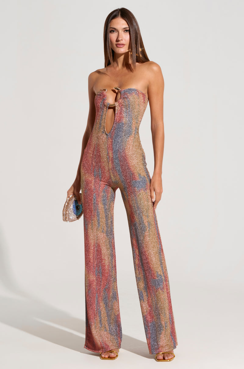 A woman stands confidently modeling the IZARA PRINTED JUMPSUIT WITH SNAKE DETAIL, a colorful and shimmering strapless piece featuring a front cutout detail. She holds a small, sparkly clutch and wears heels, her straight hair cascading down her shoulders against a plain, neutral backdrop.