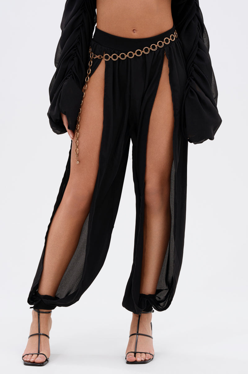 A person wears the COOL WITH ME JOGGER WITH FRONT SLITS—black, sheer, wide-leg pants with high side slits—paired with a gold chain belt and black heels. Image shows only their lower torso and legs. 1