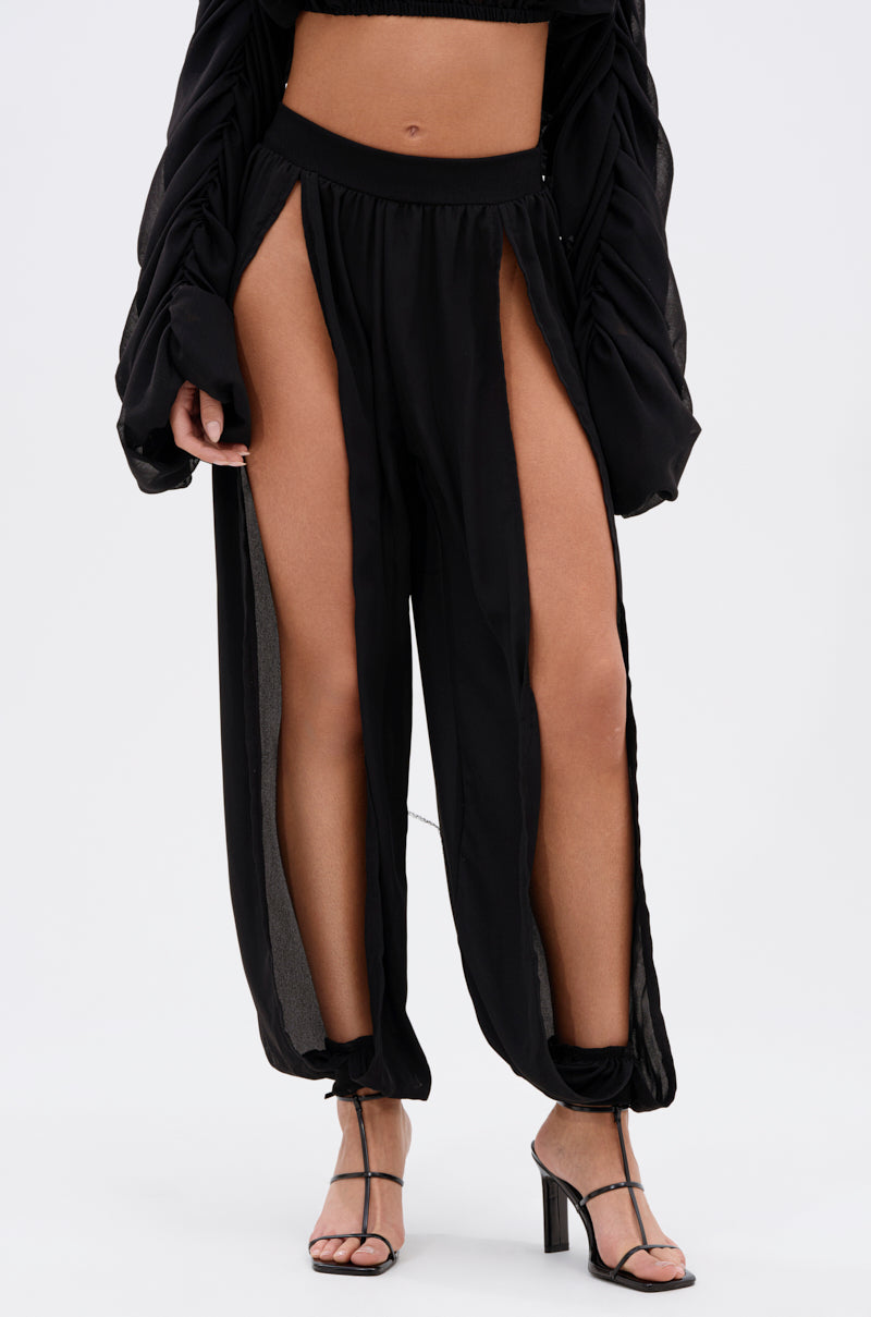 A person wears the COOL WITH ME JOGGER WITH FRONT SLITS—black joggers with open side slits and gathered ankles—paired with black high-heeled sandals and a matching black top. 2