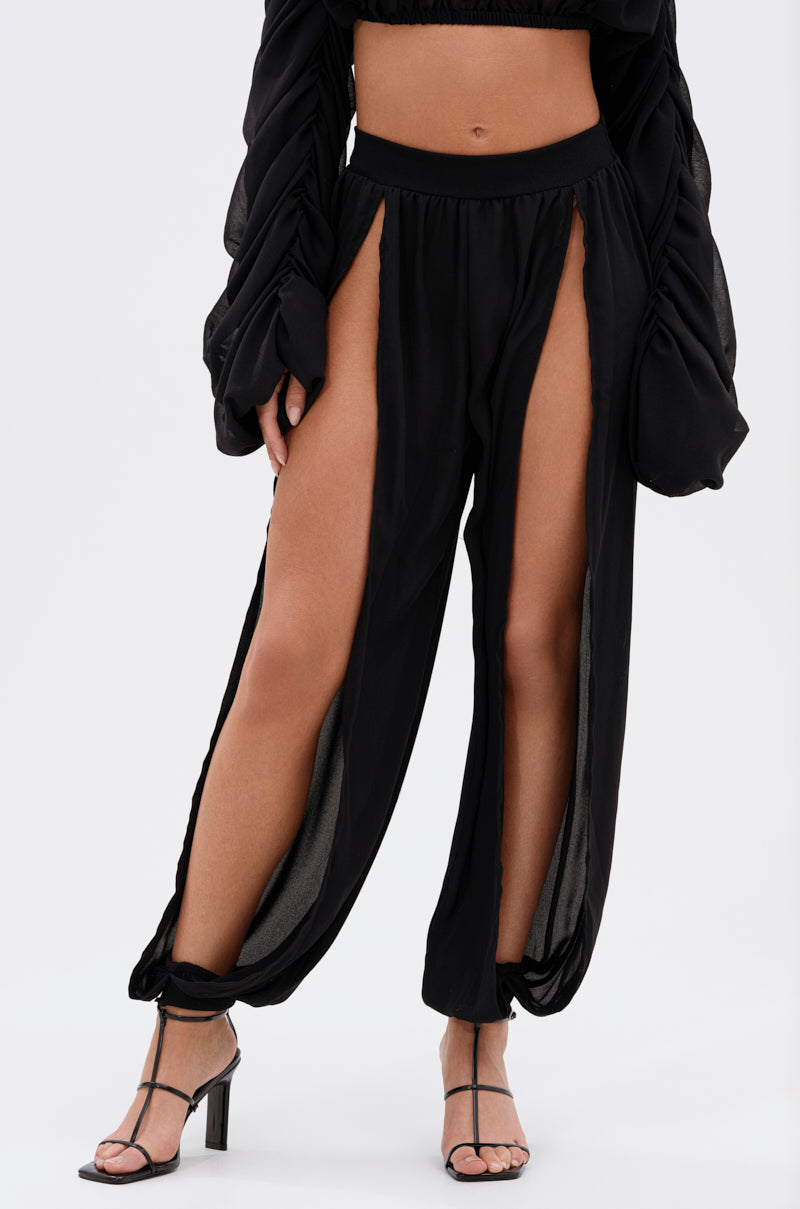 Someone wears the COOL WITH ME JOGGER WITH FRONT SLITS, featuring high front slits that reveal their legs, paired with black heeled sandals and a black long-sleeve crop top. The background is plain white. 3