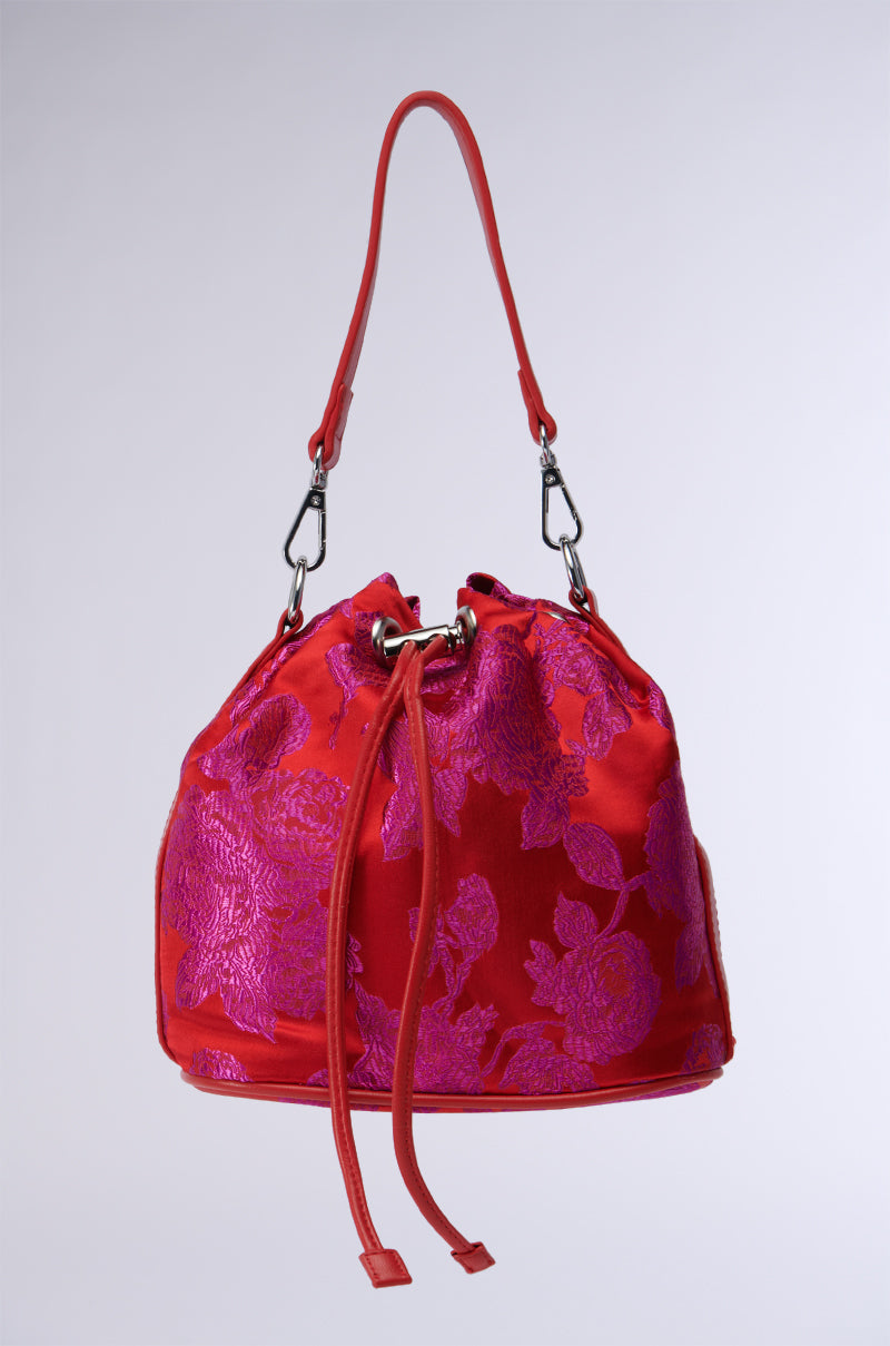 Product Description: Introducing the "ELEGANCE IS KEY BROCADE BUCKET BAG" in red, embellished with a purple floral pattern and silver hardware. It includes a drawstring closure, a single strap, and an optional detachable crossbody strap. The presentation is set against a plain white background.