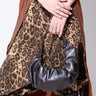 A person with crossed arms holds a small HAUTE VIBES PU PURSE. They wear a leopard-print dress with a long brown cardigan and have several silver rings. Their outfit includes black boots with black laces.