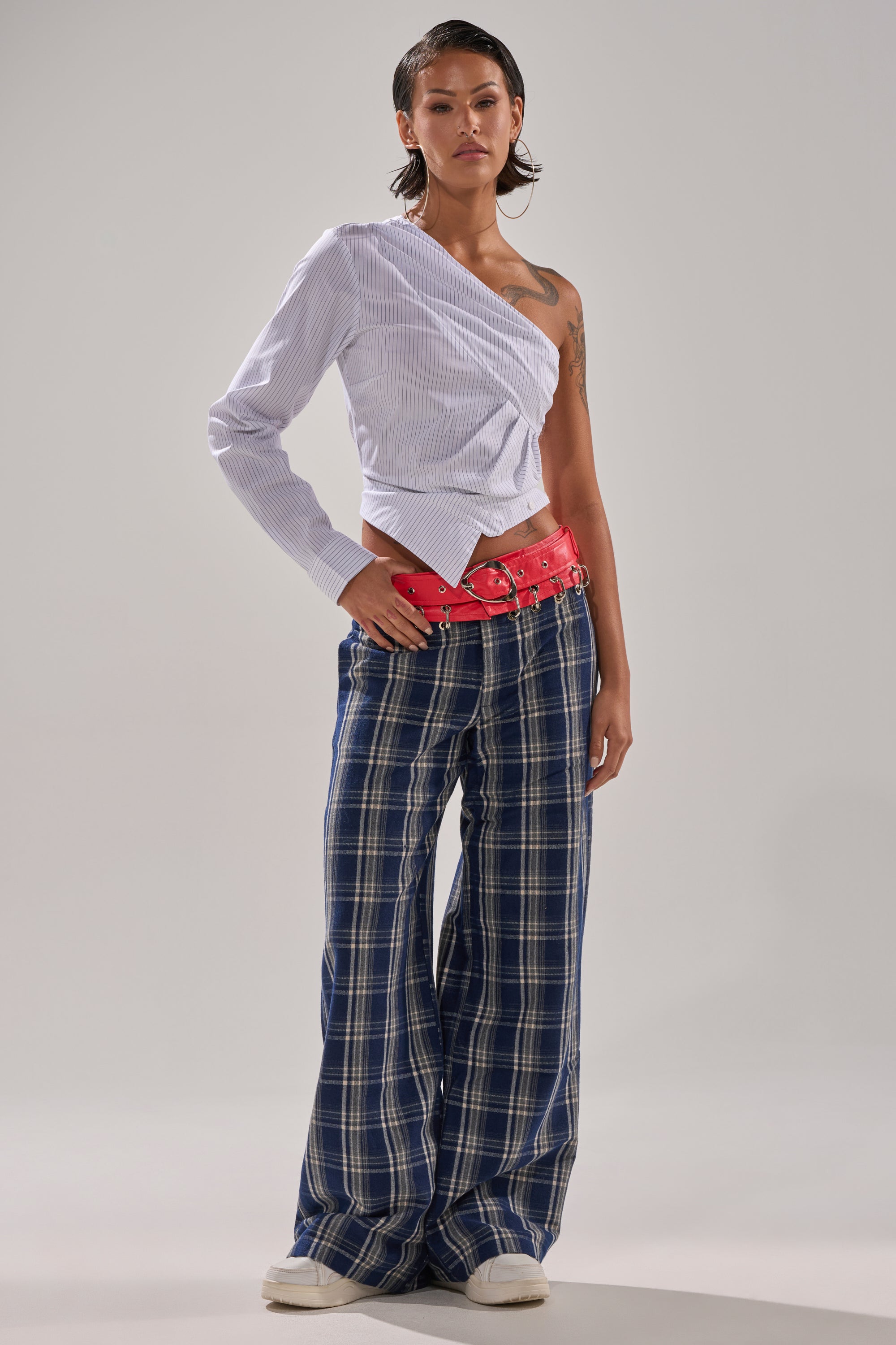 A person in a studio wears a one-sleeve white blouse, the FREE STYLE LIFE PLAID TROUSER, a red multi-buckle belt, and white shoes. They have short hair and visible tattoos on one arm.