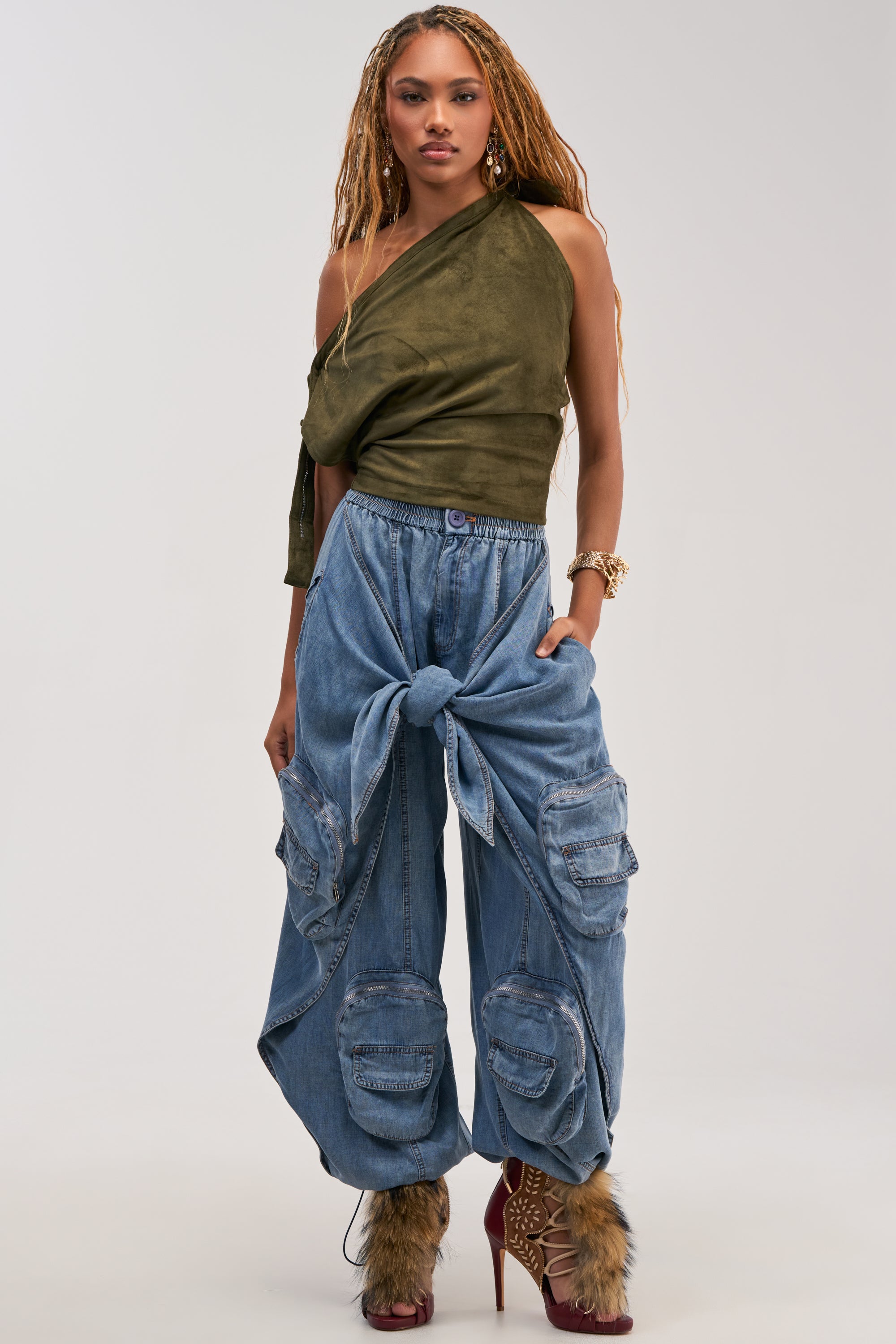 A woman stands against a plain background wearing an olive green one-shoulder top and the DO THE MOST TIE FRONT PANT IN DENIM, which features a wide leg cargo design with multiple pockets and a front tie detail. She completes the look with red lace-up heels with fur accents and jewelry.