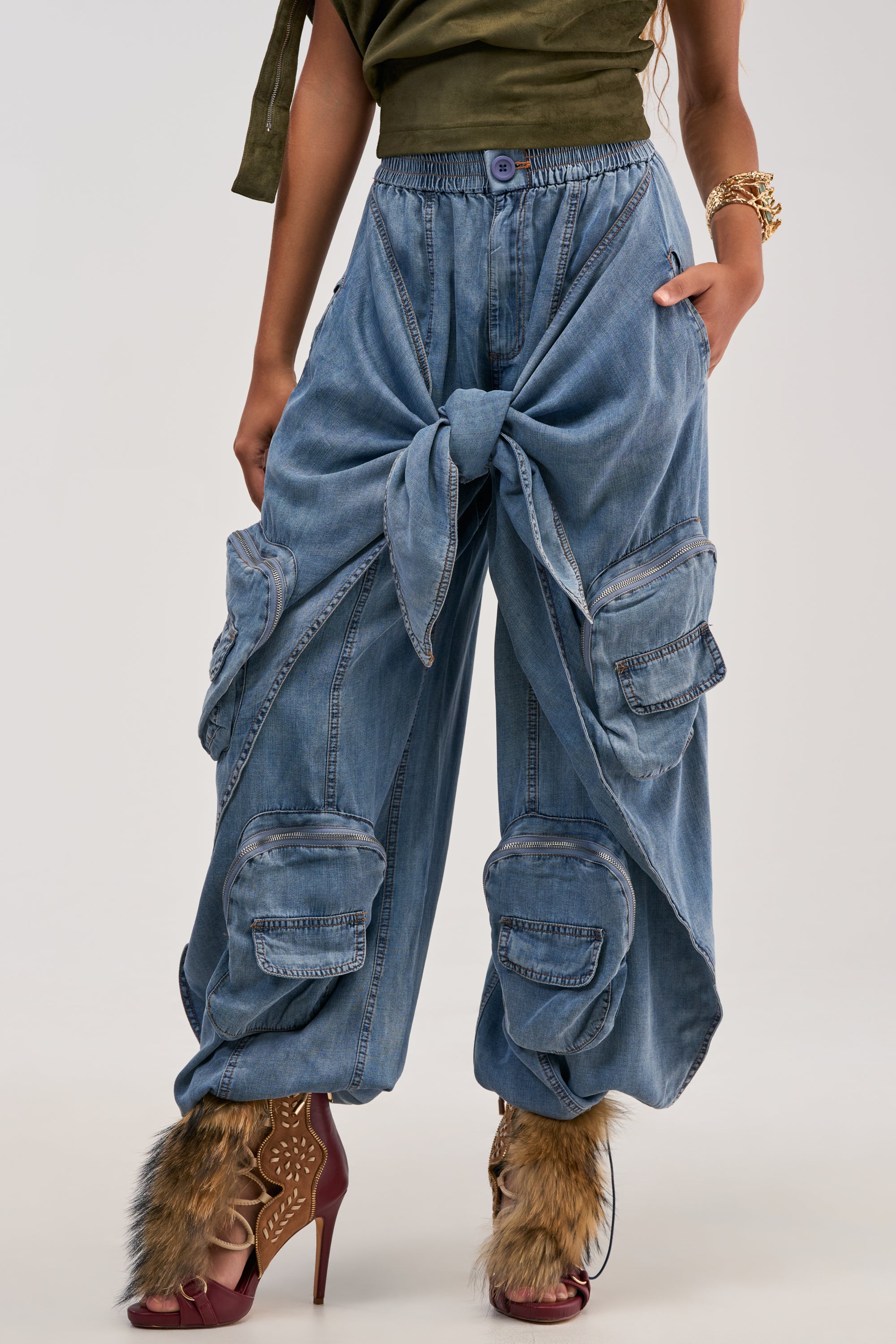 A person showcasing the lower half of their body wears the DO THE MOST TIE FRONT PANT IN DENIM, featuring a distinctive oversized front knot and multiple cargo pockets; the look is completed with open-toe heels adorned with faux fur for an edgy streetwear style.