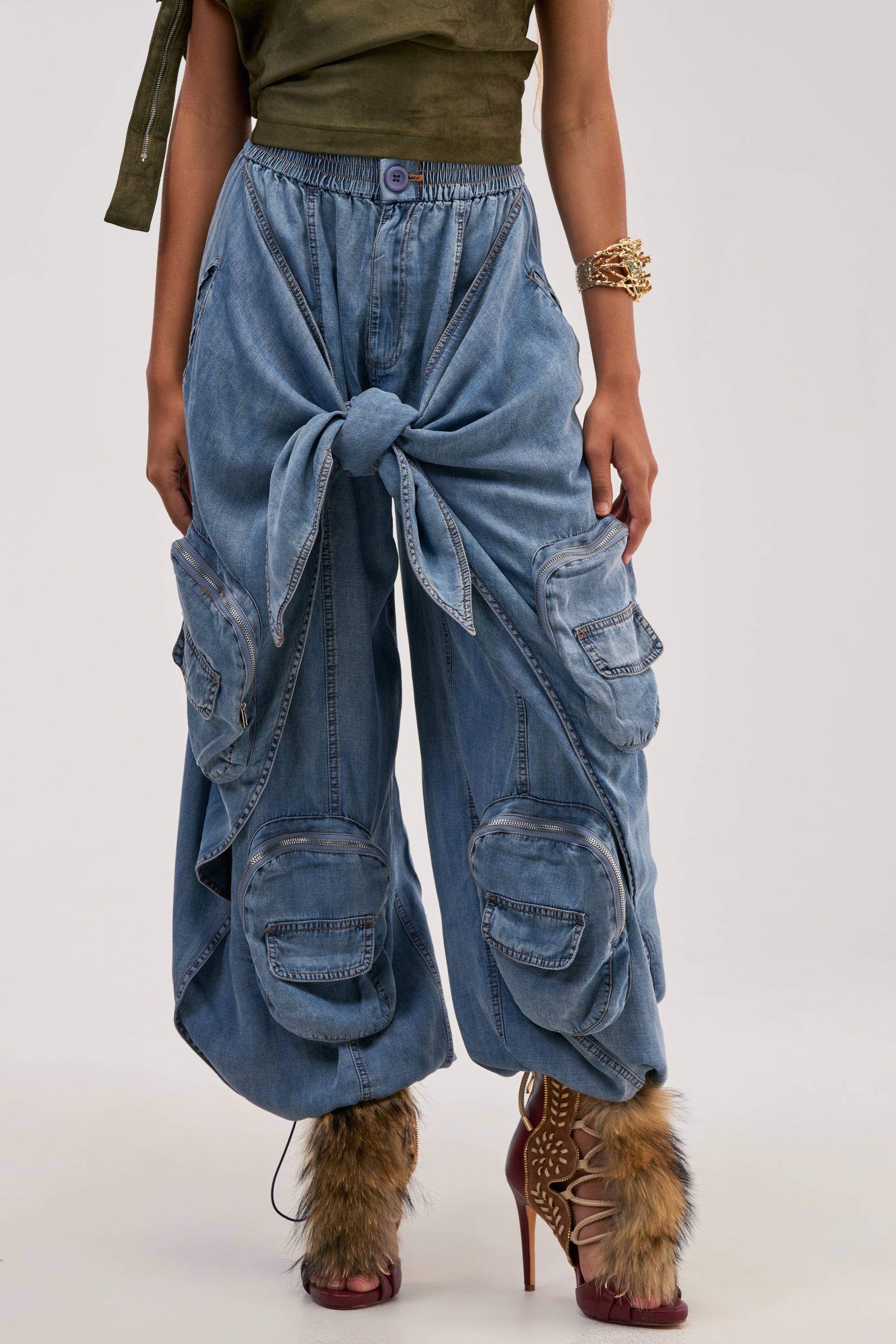 Wearing the “DO THE MOST TIE FRONT PANT IN DENIM”—wide-leg blue denim cargo pants featuring large pockets and an oversized front knot—this look is paired with brown furry high-heeled shoes, a green top, and bracelets. The outfit exudes an effortlessly cool streetwear vibe against a plain, light background.