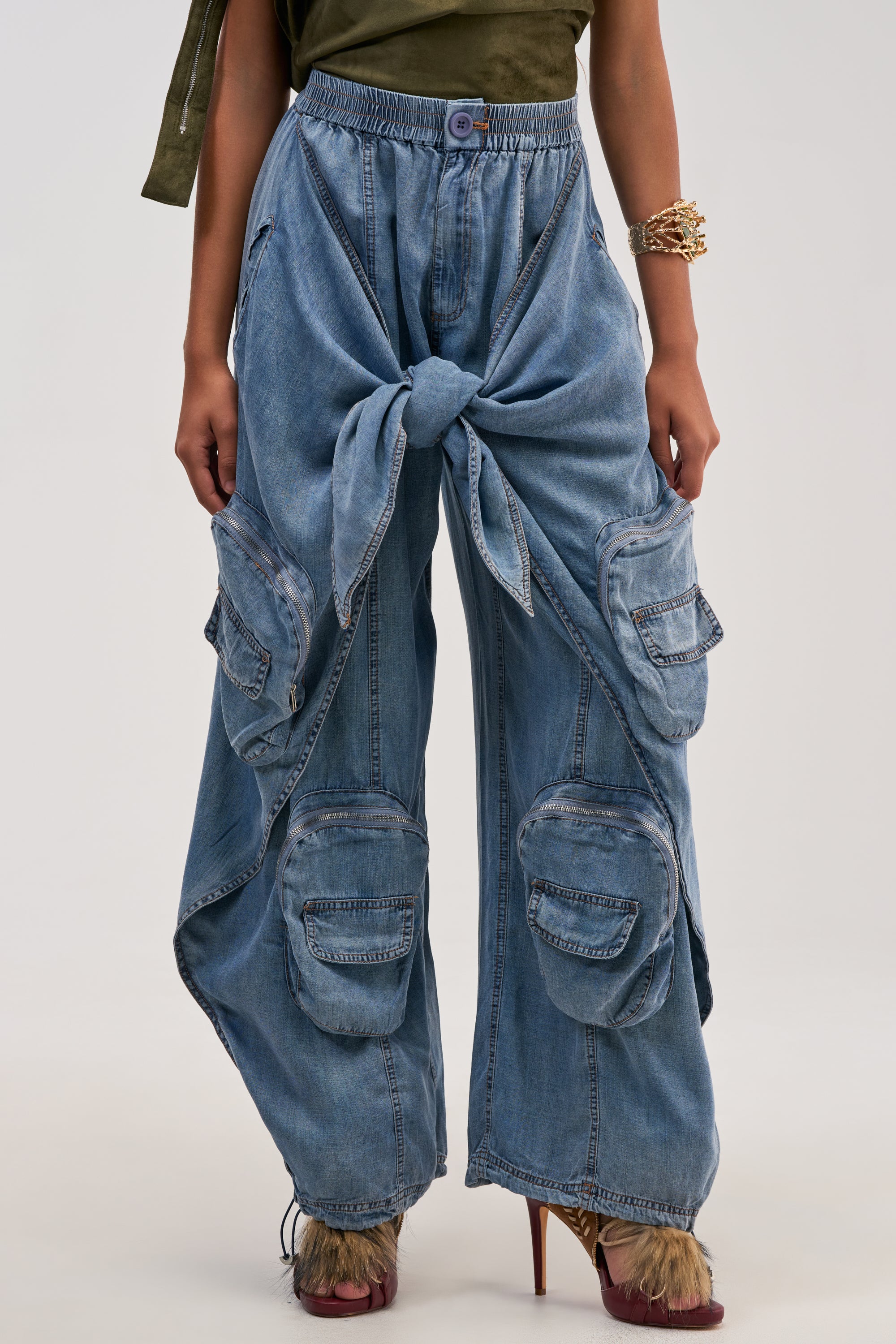 Wearing the DO THE MOST TIE FRONT PANT IN DENIM—featuring wide-leg denim, a dramatic front knot, and multiple cargo pockets—paired with furry high-heeled shoes creates a bold streetwear vibe. The look is completed with a green top and gold bracelet, while the model’s face remains unseen.