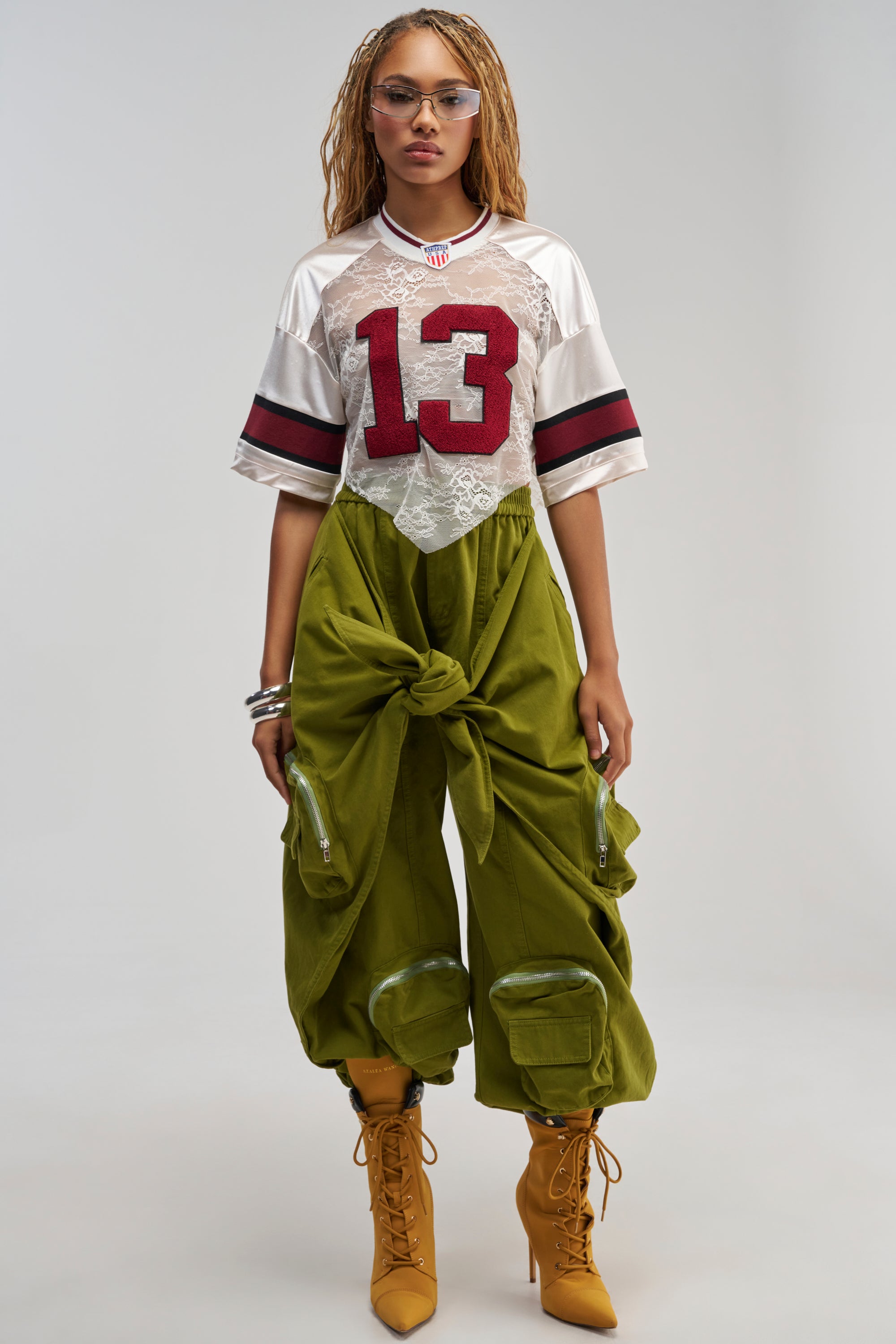 A woman stands against a plain background wearing the DO THE MOST TIE FRONT JOGGER IN OLIVE paired with a sporty white and red lace jersey, yellow lace-up boots, clear glasses, and bracelets.