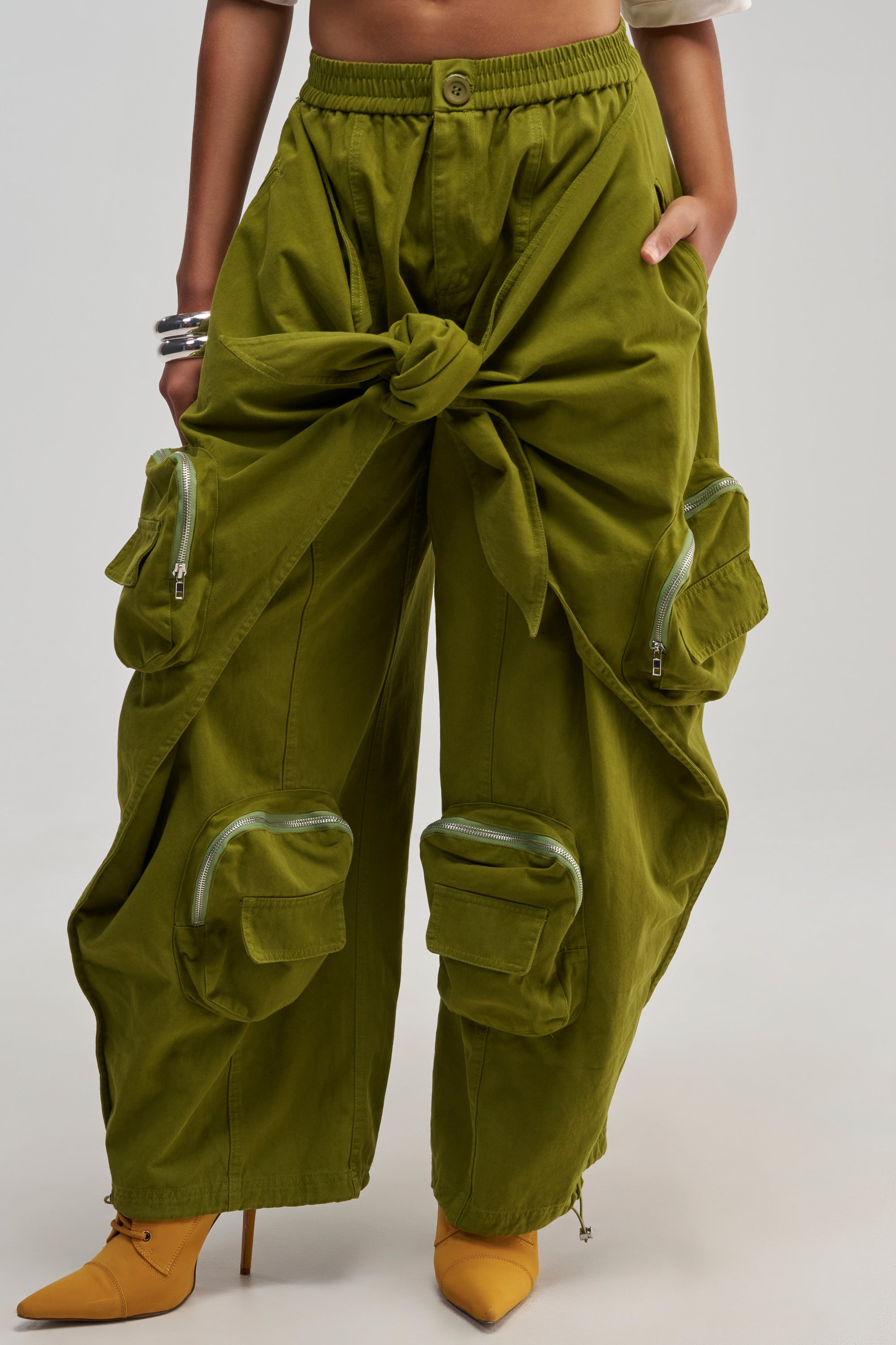 Wearing the DO THE MOST TIE FRONT JOGGER IN OLIVE—featuring oversized pockets, zippers, and a prominent front tie—this bold streetwear-inspired outfit is styled with yellow pointed heels and silver bracelets.