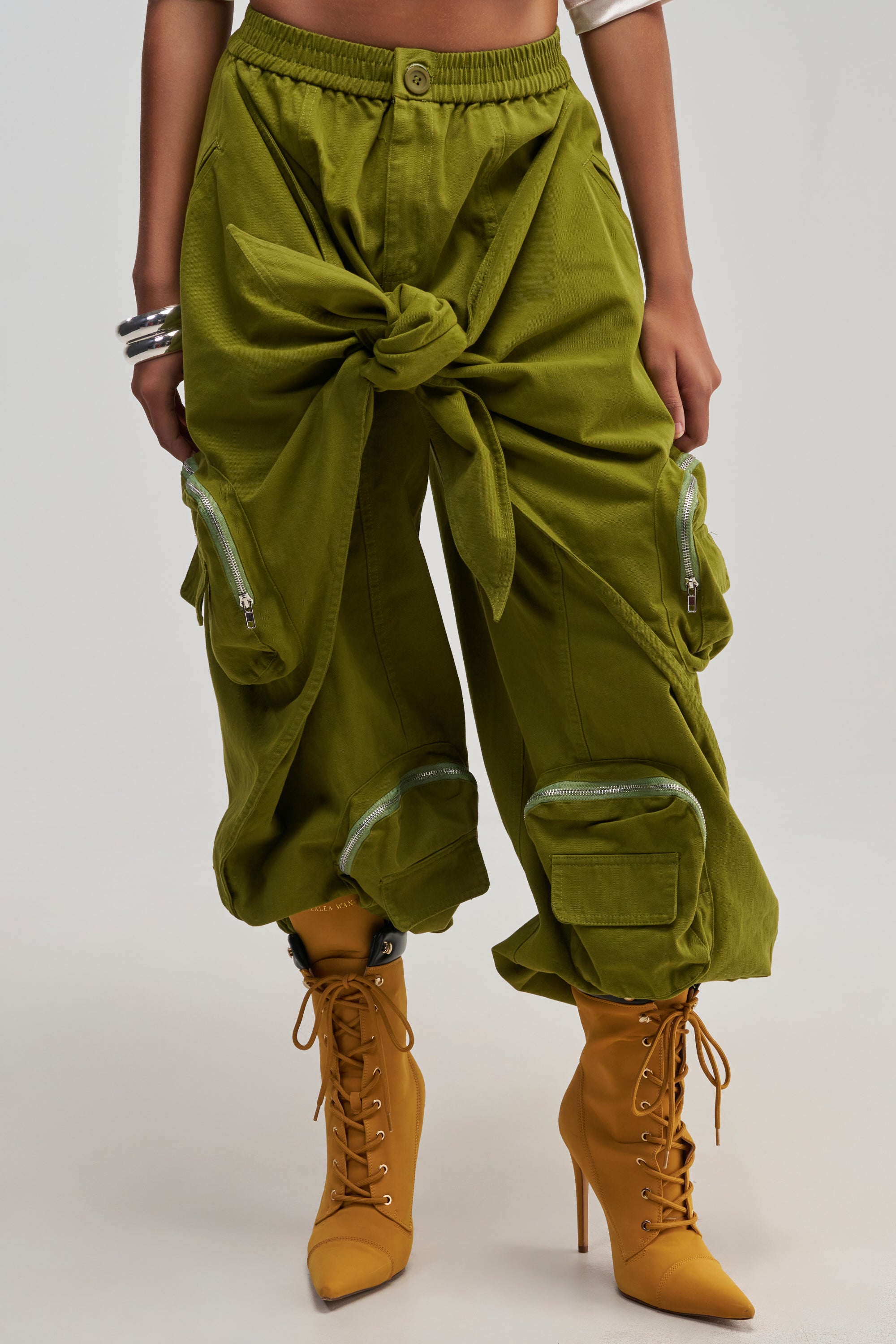 A person showcases the lower half of their outfit, wearing the DO THE MOST TIE FRONT JOGGER IN OLIVE—a pair of streetwear-inspired cargo pants with zippered pockets, a relaxed fit, and a distinctive large tie at the front—paired with tan lace-up heeled boots.