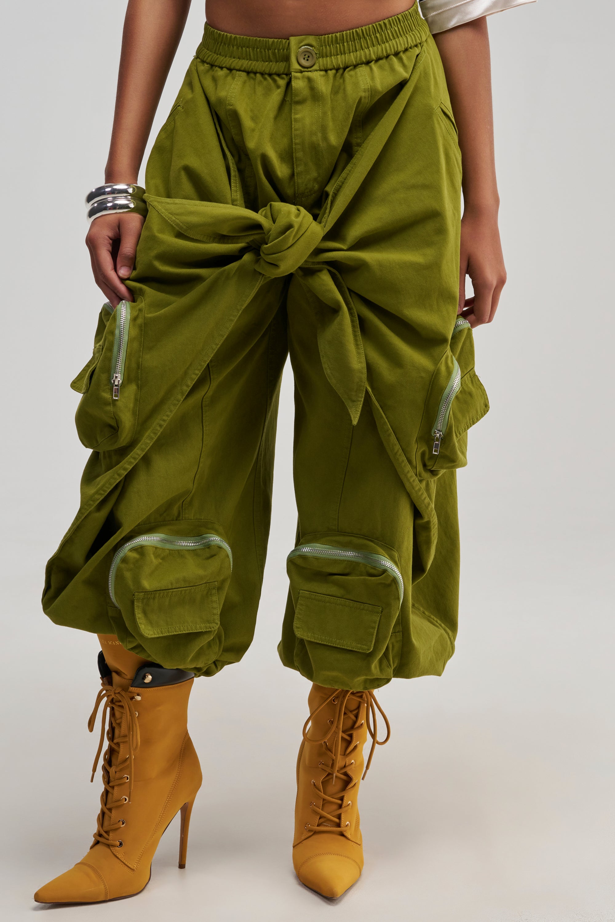 Wearing the DO THE MOST TIE FRONT JOGGER IN OLIVE with a knotted waist and roomy pockets, styled with mustard yellow heeled lace-up boots and silver bangles. This streetwear-inspired outfit showcases only the lower torso and legs.