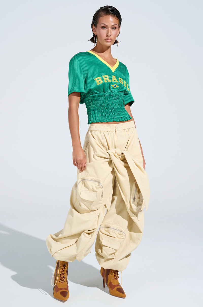 A woman wears a green "Brazil" V-neck top and beige DO THE MOST TIE FRONT PANT, high-rise and tied at the waist. She pairs this streetwear look with tan lace-up boots, standing confidently against a plain light background.