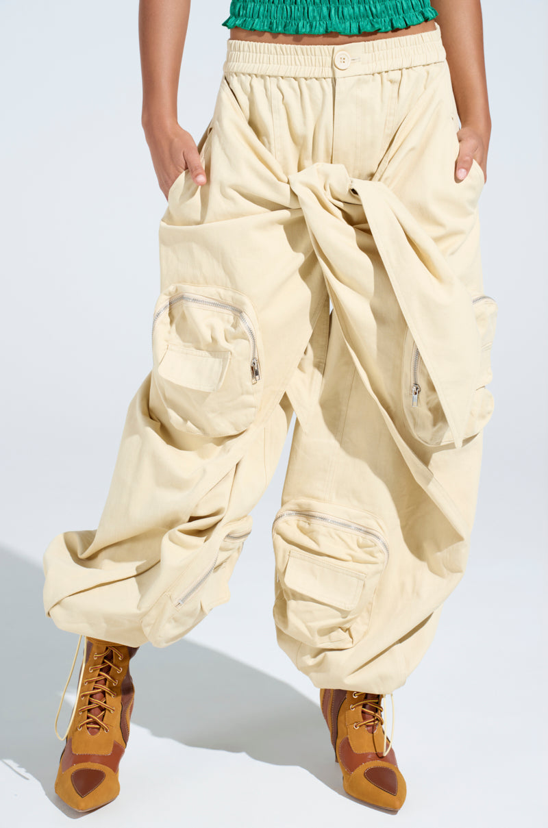 Person wearing the DO THE MOST TIE FRONT PANT—oversized, cream cargo pants with a high-rise fit and statement pockets—paired with tan lace-up boots and a green top. Photo cropped at the torso, spotlighting pants and boots in a streetwear style.
