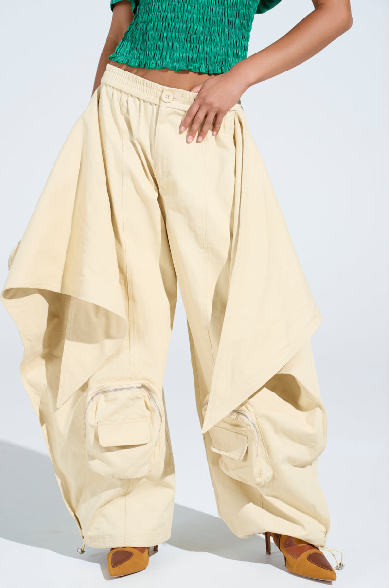 A person wears DO THE MOST TIE FRONT PANT in a wide cream high-rise fit with large draped panels and pockets, paired with a green textured top and brown pointed-toe heels, standing against a plain background.