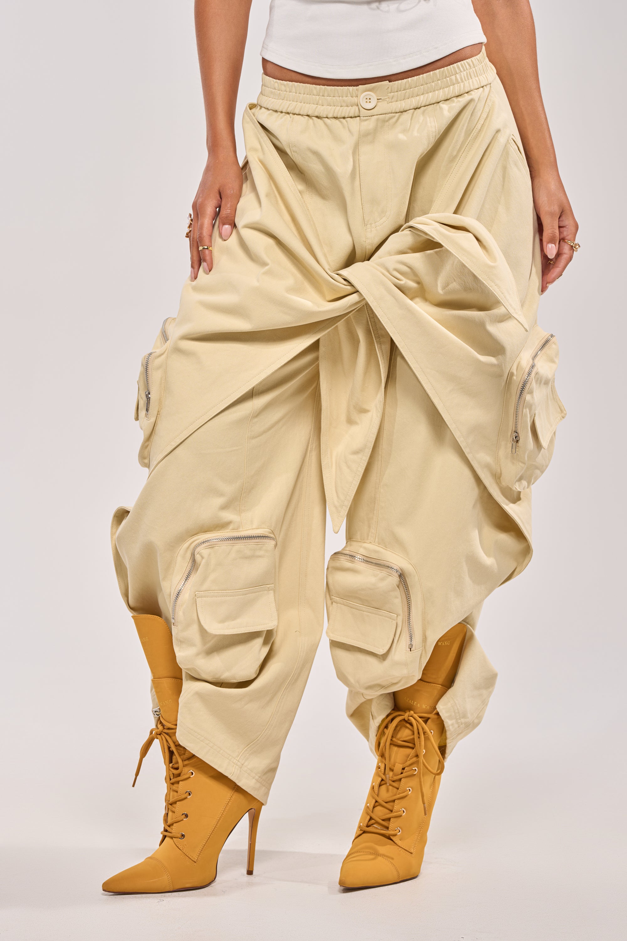 A person models the DO THE MOST TIE FRONT PANT—oversized khaki cargo pants with exaggerated folds—paired with beige lace-up heeled boots, shown from the waist down against a light background for a streetwear vibe.