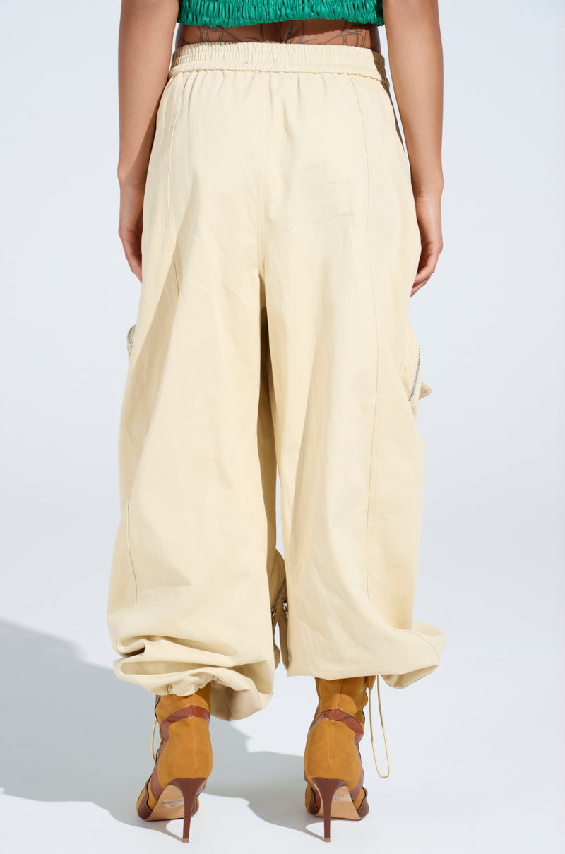 Person wearing DO THE MOST TIE FRONT PANT in a high-rise fit, shown from the back with tan heeled sandals against a plain light background. The streetwear-inspired beige pants are loosely bunched at the ankles.