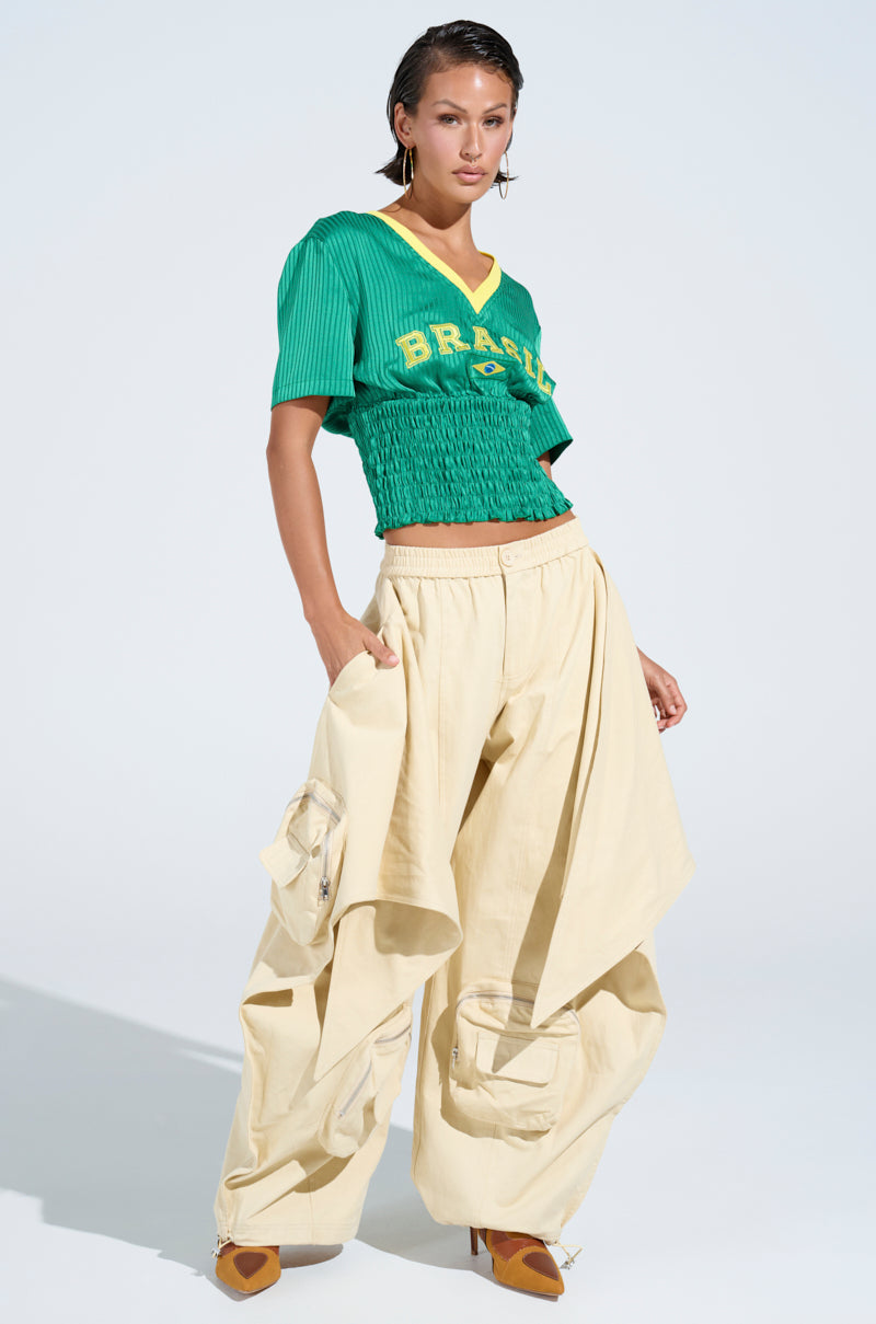 A woman poses confidently in a green "Brazil" crop top and the DO THE MOST TIE FRONT PANT with oversized pockets, paired with brown heels, creating a bold streetwear look against a plain light background.