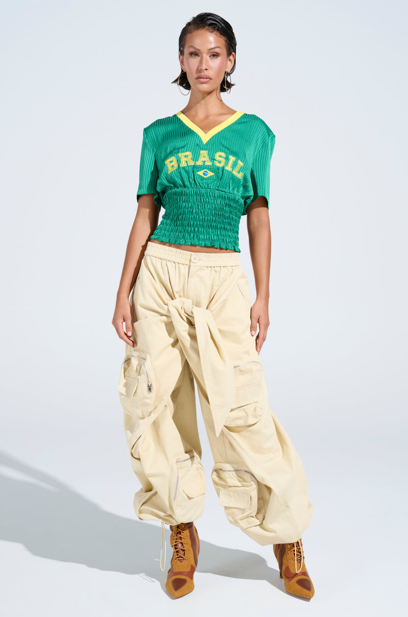 A woman poses in a green "Brasil" shirt, DO THE MOST TIE FRONT PANT cargo pants with a cinched waist, and brown lace-up boots—showcasing effortless streetwear style against a plain light background.