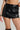 A person wearing the THINK ABOUT IT MINI SKORT, featuring curve-hugging design and metallic zipper details, accessorized with a silver bracelet on their left wrist. They are also donning black knee-high boots, visible just at the top of the image, set against a plain gray background.