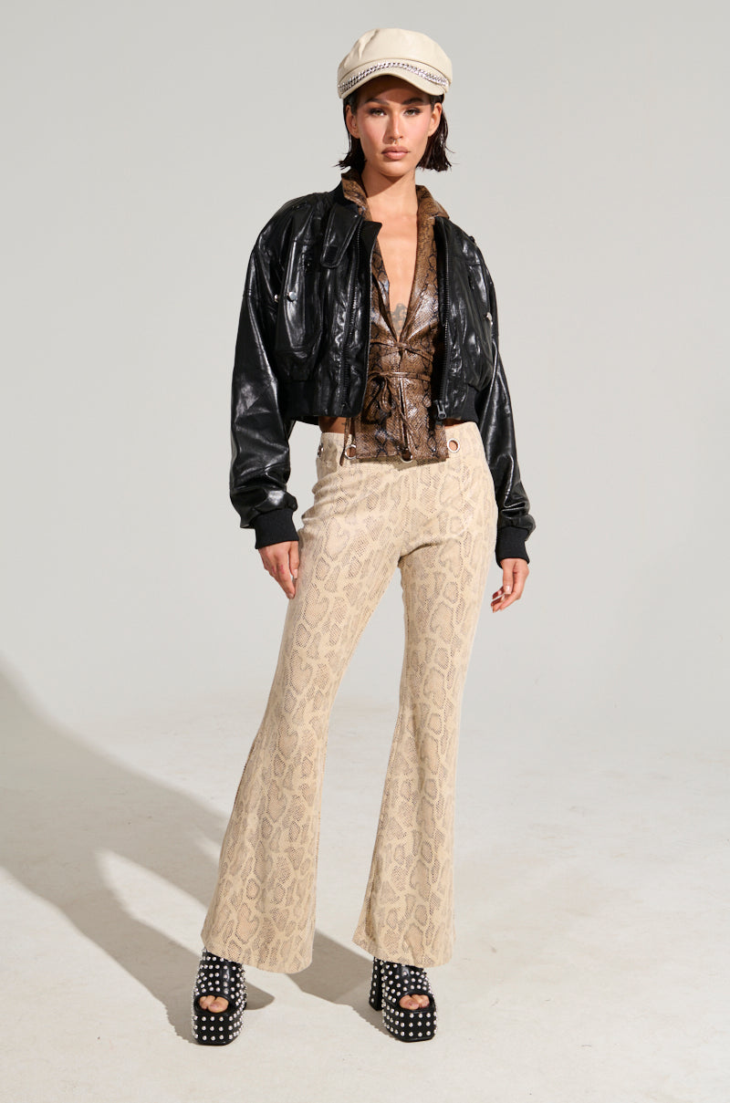 A person exudes confidence modeling a trendy ensemble that features a black leather jacket, the SNAKE CHARMER PANT with an embossed snakeskin print, and studded platform sandals. They top off the look with a white newsboy cap, posing stylishly against a plain background.