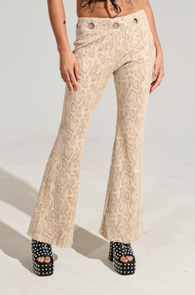 Dressed in their SNAKE CHARMER PANT featuring a beige embossed snakeskin print and detailed with silver grommets at the waist, a person confidently showcases their style. They complete the ensemble with black high-heeled sandals adorned with silver studs.
