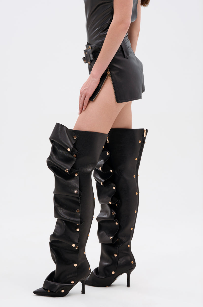 A person wearing the TOO HOT FOR YOU FAUX LEATHER HOT SHORT and high-heeled, over-the-knee black boots with ruffled details and gold-tone hardware, standing against a plain white background.