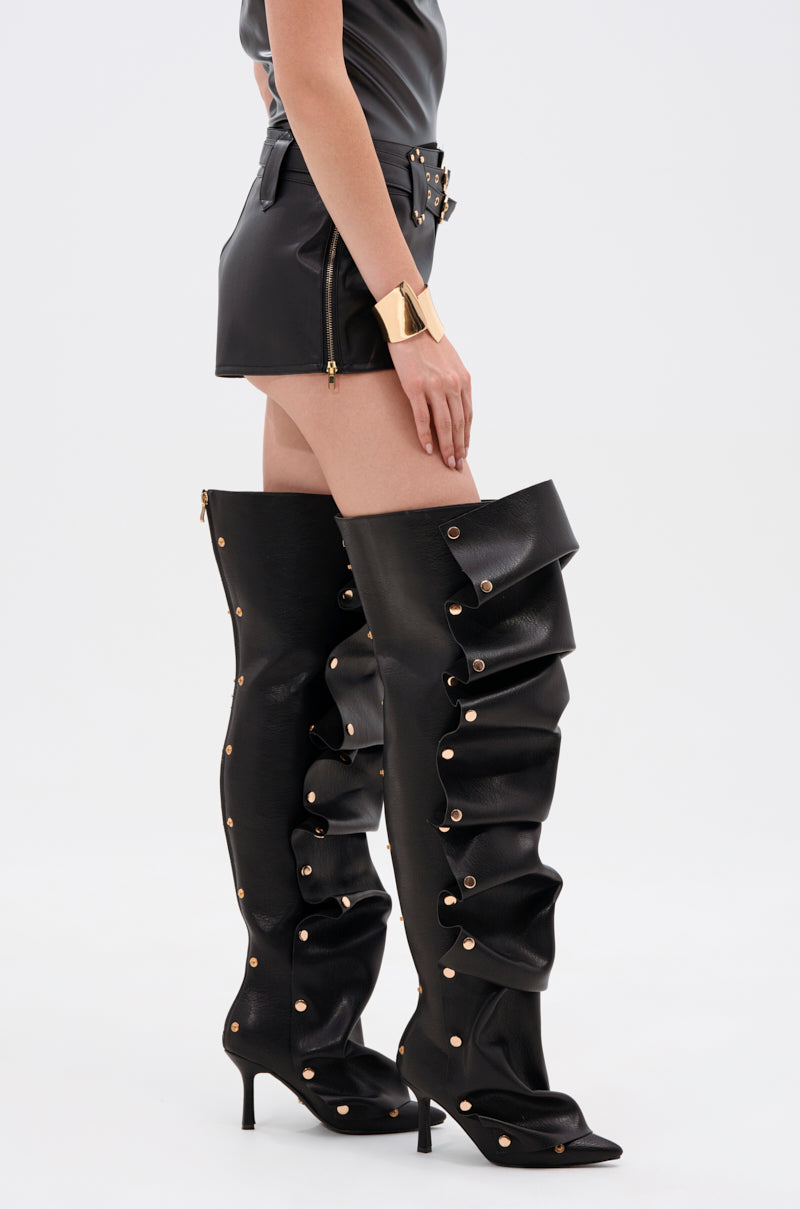 Wearing the TOO HOT FOR YOU FAUX LEATHER HOT SHORT with a black top and dramatic black thigh-high boots featuring gold-tone hardware, pointed toes, and stiletto heels. The look is finished with a gold cuff bracelet.