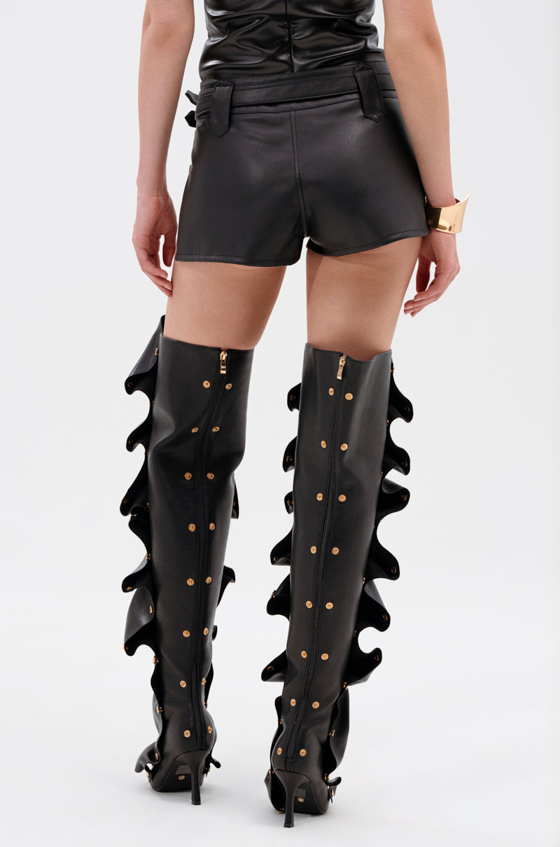 A person models the TOO HOT FOR YOU FAUX LEATHER HOT SHORT and thigh-high black boots with gold-tone hardware, gold studs, and ruffled edges, photographed from the back against a plain background.