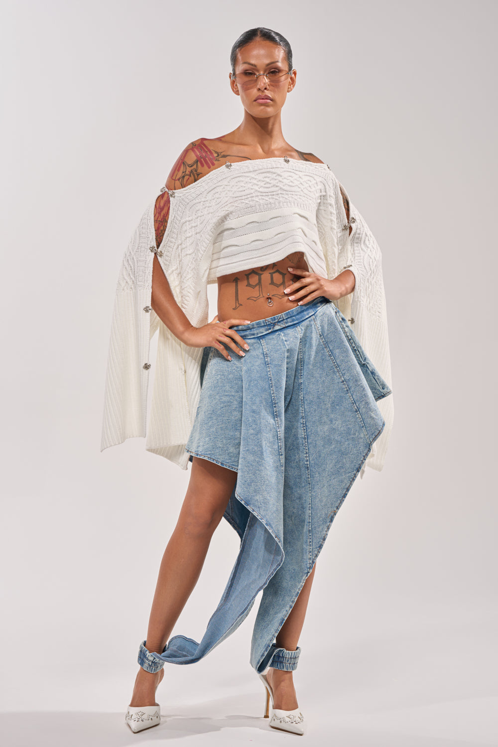 A woman exudes street style in a white off-shoulder knit top and COOLEST EVER DENIM high-rise cuffed jeans. She pairs them with sunglasses, visible tattoos, and white heels, posing confidently against a plain backdrop. 0