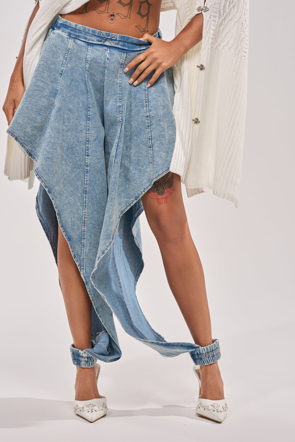 A person wears the COOLEST EVER DENIM—unique, asymmetrical, light blue street-style pants with pointed hems and ankle fastenings—paired with white heeled shoes and a white textured cardigan draped over their shoulders. 2