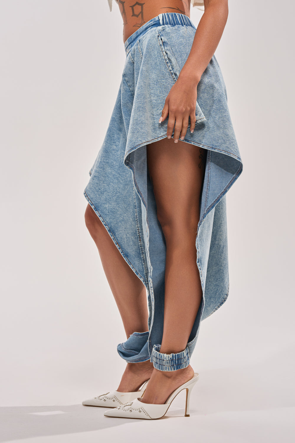 Someone stands against a plain background wearing COOLEST EVER DENIM high-rise, asymmetrical hem jeans, white high-heeled shoes, and displays a side tattoo. 4