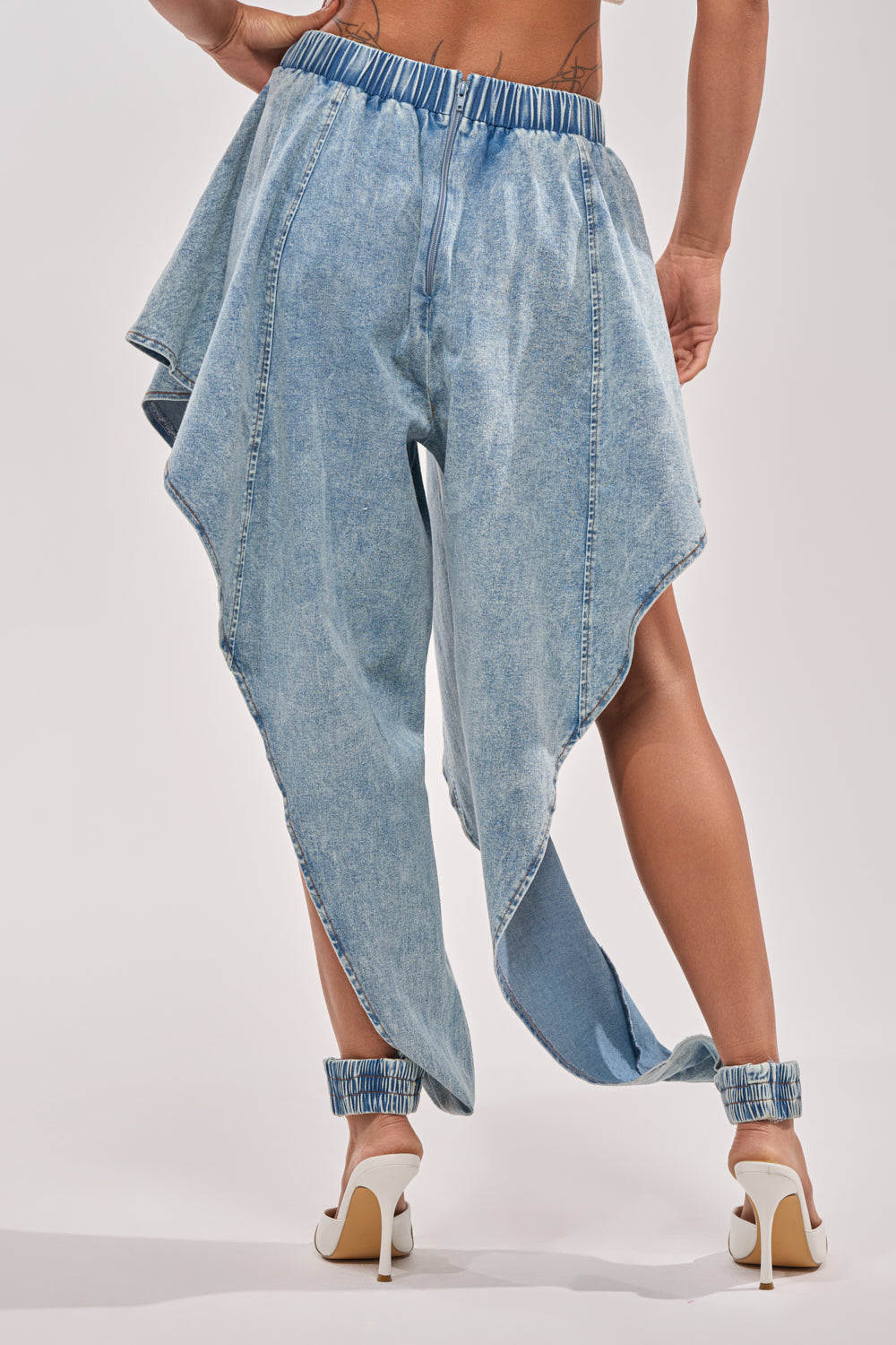 Model showcases the COOLEST EVER DENIM—light blue, high-rise jeans with asymmetrical draped legs and gathered elastic cuffs—paired with white heels, viewed from the back against a plain light background. 5