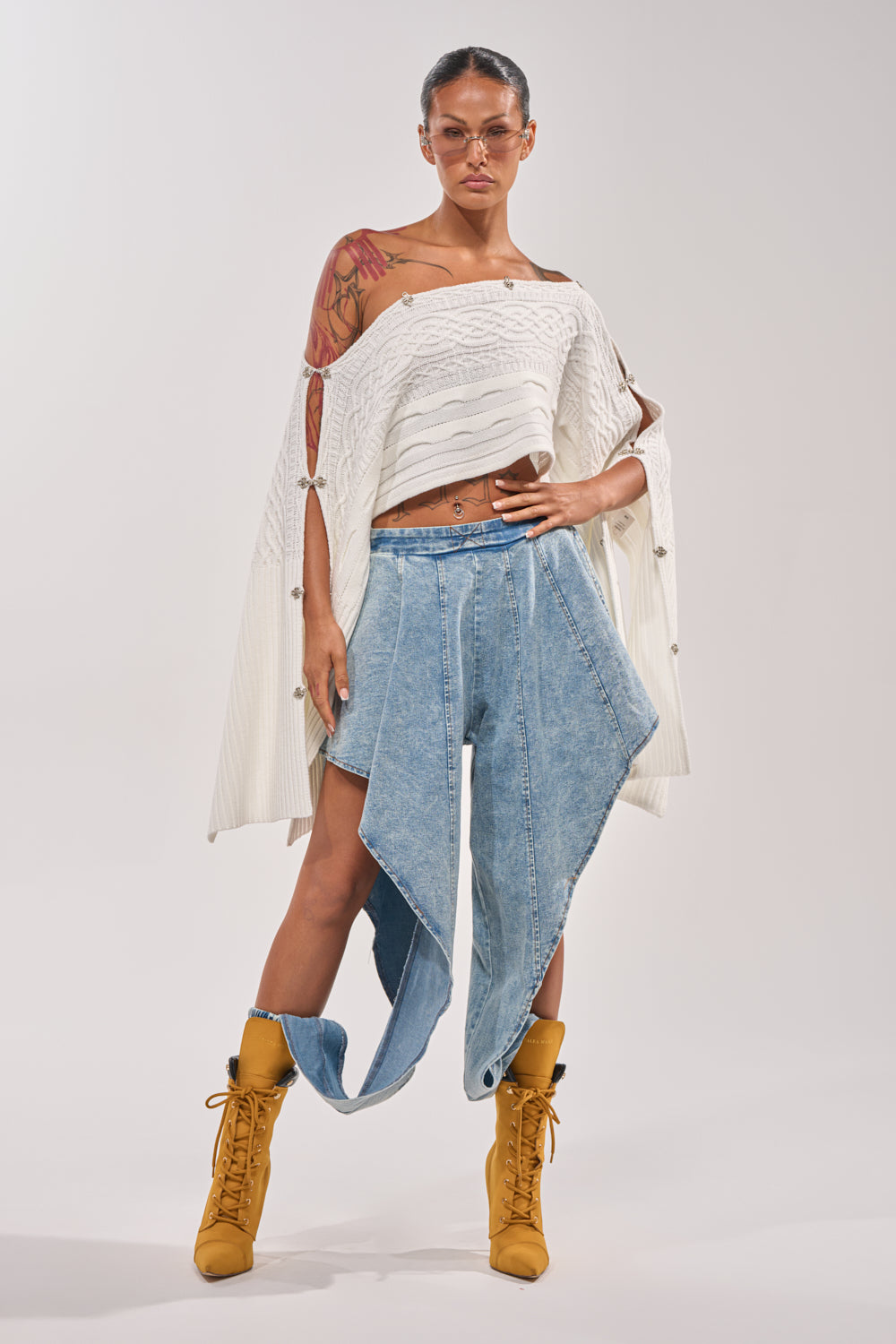 A woman with tattoos wears a white off-shoulder sweater, yellow lace-up boots, clear glasses, and COOLEST EVER DENIM high-rise jeans with asymmetrical layered panels. She stands confidently against a plain background. 6