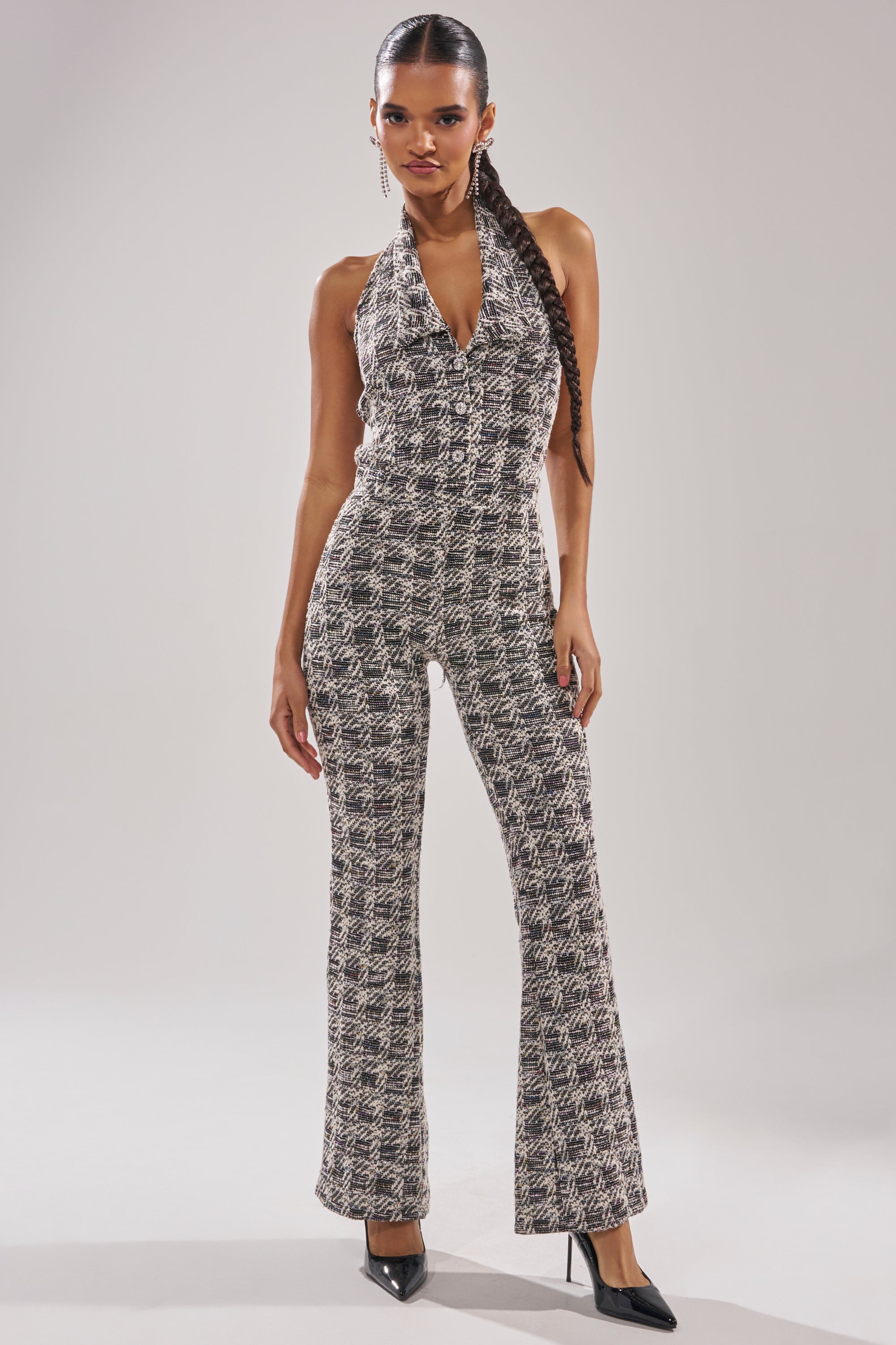 A woman stands confidently in the CHECKMATE SLEEVELESS JUMPSUIT with flared legs and pointed black heels. Her hair is styled in a sleek braid as she poses against a plain, light-colored background. 0