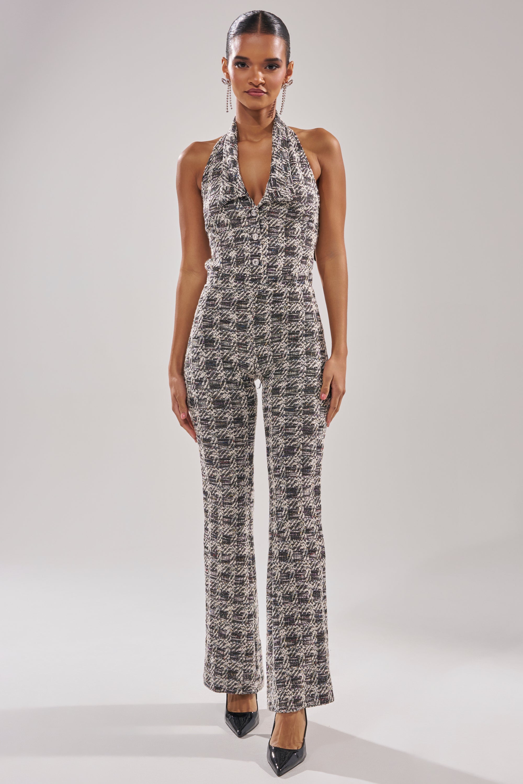 A woman faces forward in the CHECKMATE SLEEVELESS JUMPSUIT, featuring a houndstooth print, halter neckline, and flared legs, styled with black pointed-toe heels against a plain light background. 2