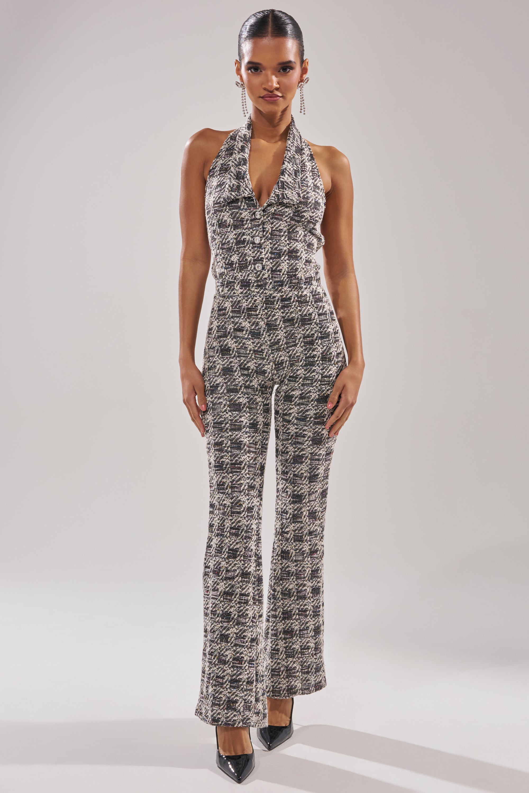 A woman stands against a plain light background wearing the CHECKMATE SLEEVELESS JUMPSUIT with a halter neck, paired with black high heels and long dangling earrings. 3