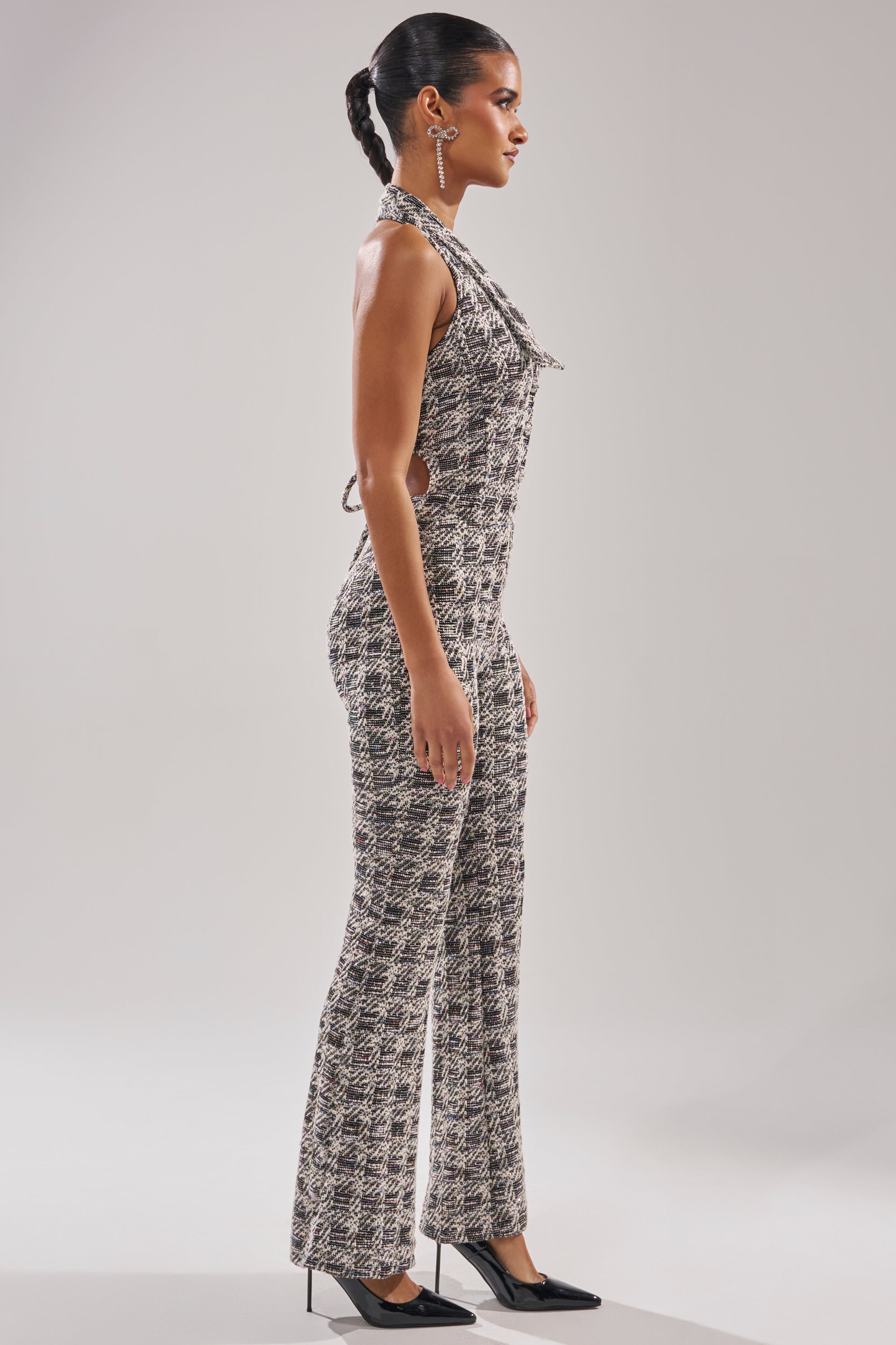 A woman in profile wears the CHECKMATE SLEEVELESS JUMPSUIT with flared legs and black heels. Her hair is pulled back, she's styled with ear cuffs, and she stands against a plain light background. 4