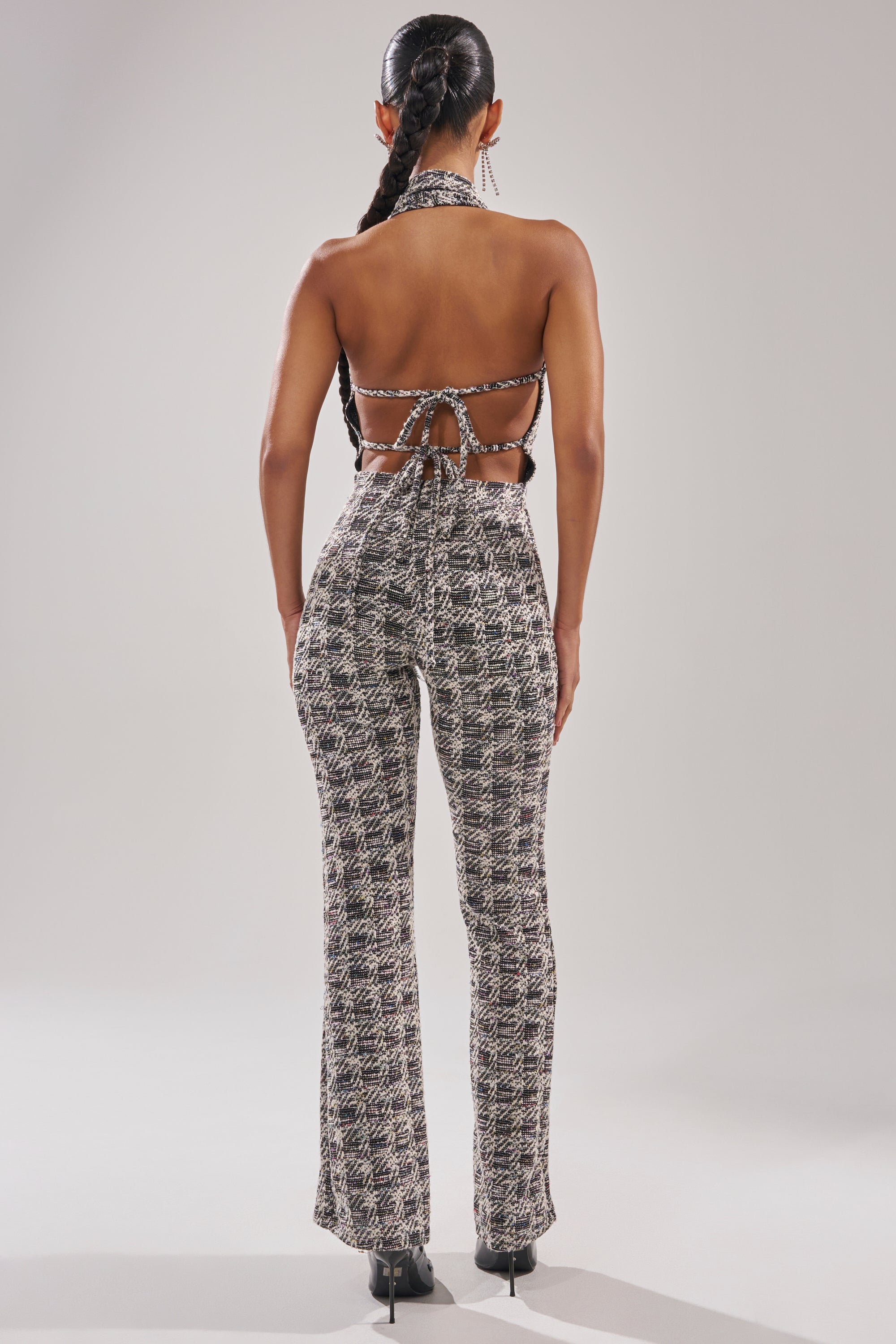 A woman with a long braid stands with her back to the camera, wearing the CHECKMATE SLEEVELESS JUMPSUIT—a houndstooth design featuring a halter neckline, open back, and high-waisted flared legs for a bold, textured look. 5