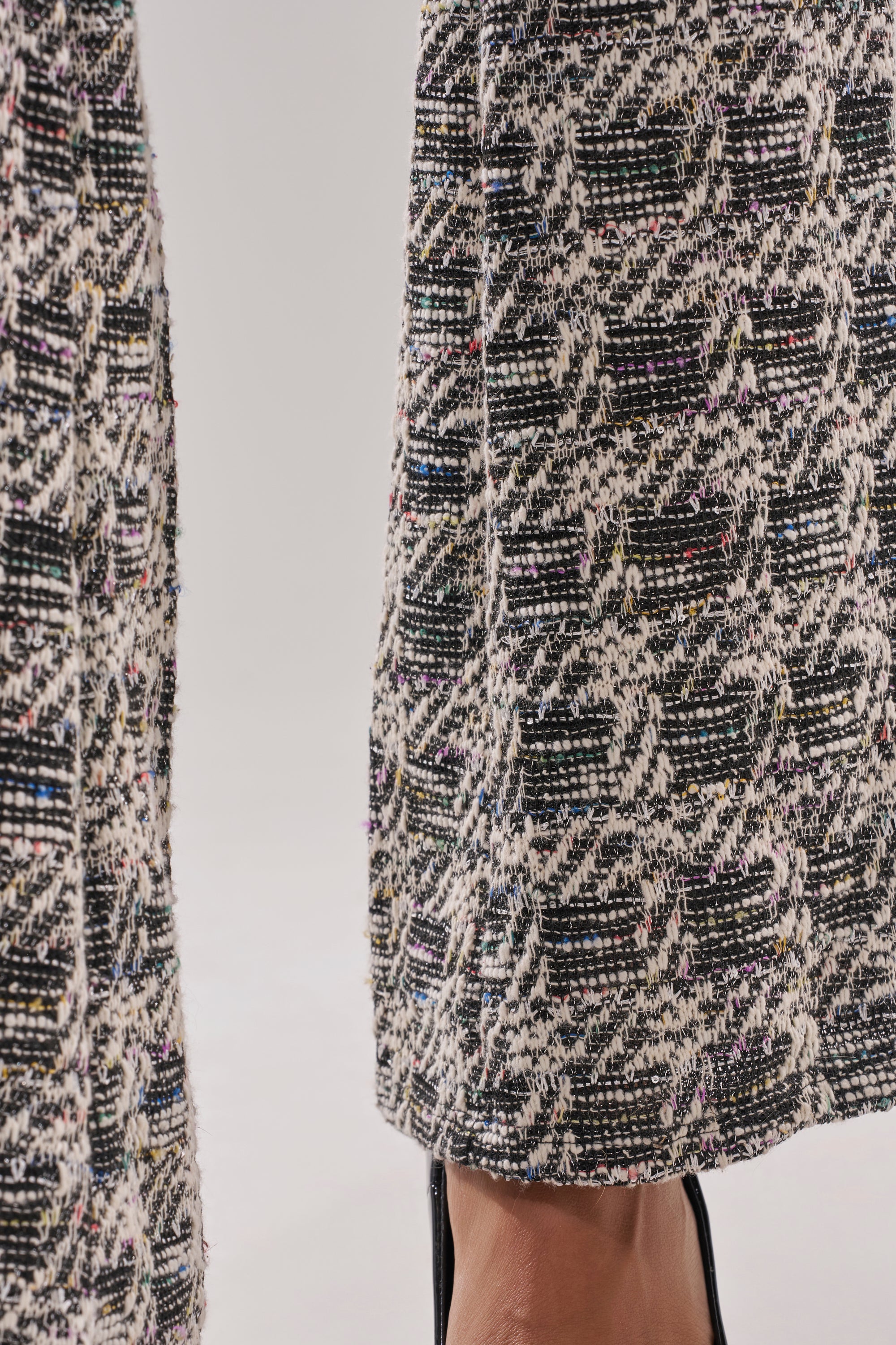 Close-up of a person wearing the CHECKMATE SLEEVELESS JUMPSUIT featuring black and white houndstooth with multicolored threads, slightly flared hem, and black heeled shoes partially visible. 6