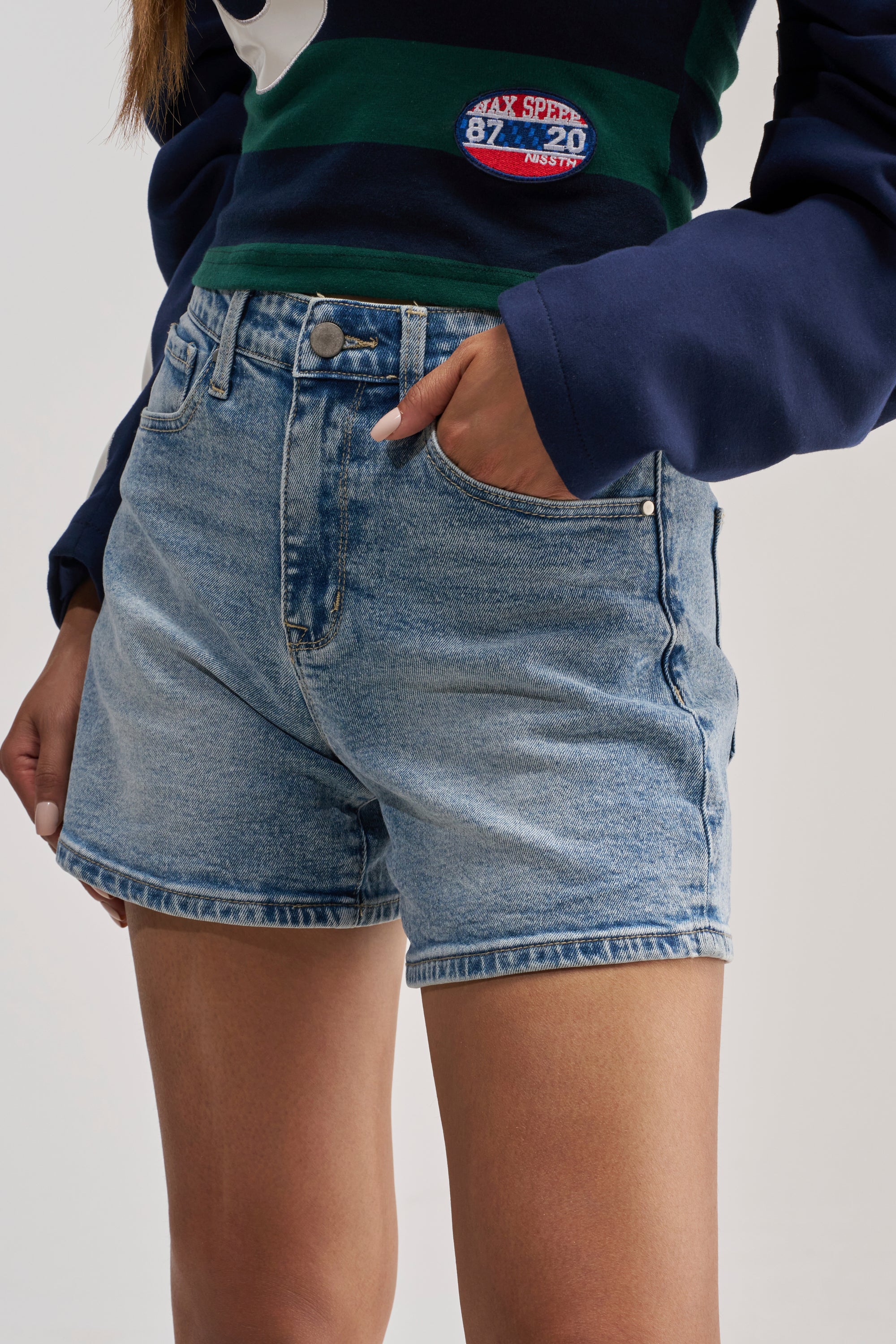 A person wearing THE BEST DENIM SHORT—a medium wash, high-rise denim short—paired with a dark long-sleeve striped top, one hand casually tucked in the pocket, shown from waist to mid-thigh against a plain background.