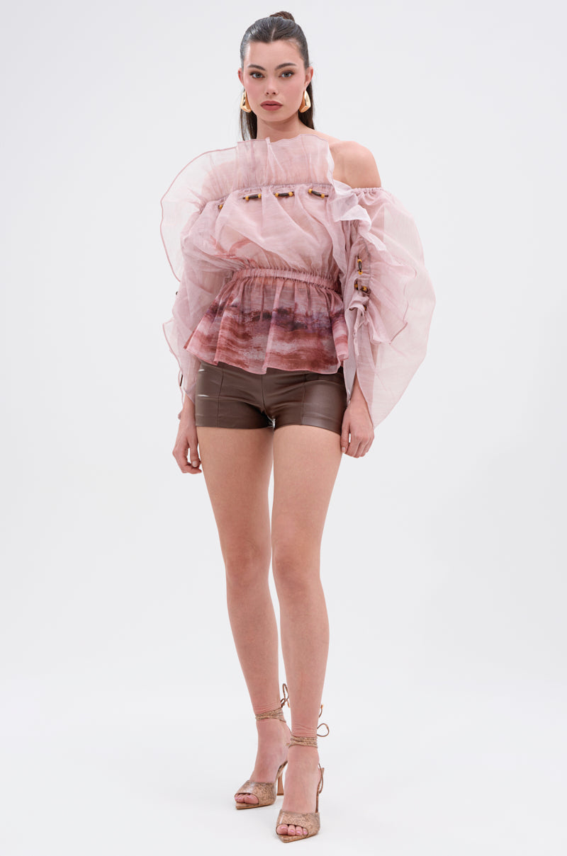 A woman poses against a white background wearing CLUB HIGH WAISTED PLEATHER SHORTS with a sheer pink ruffled top, voluminous sleeves, strappy nude heels, and her hair pulled back. 5
