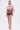 A woman poses against a white background wearing CLUB HIGH WAISTED PLEATHER SHORTS with a sheer pink ruffled top, voluminous sleeves, strappy nude heels, and her hair pulled back.