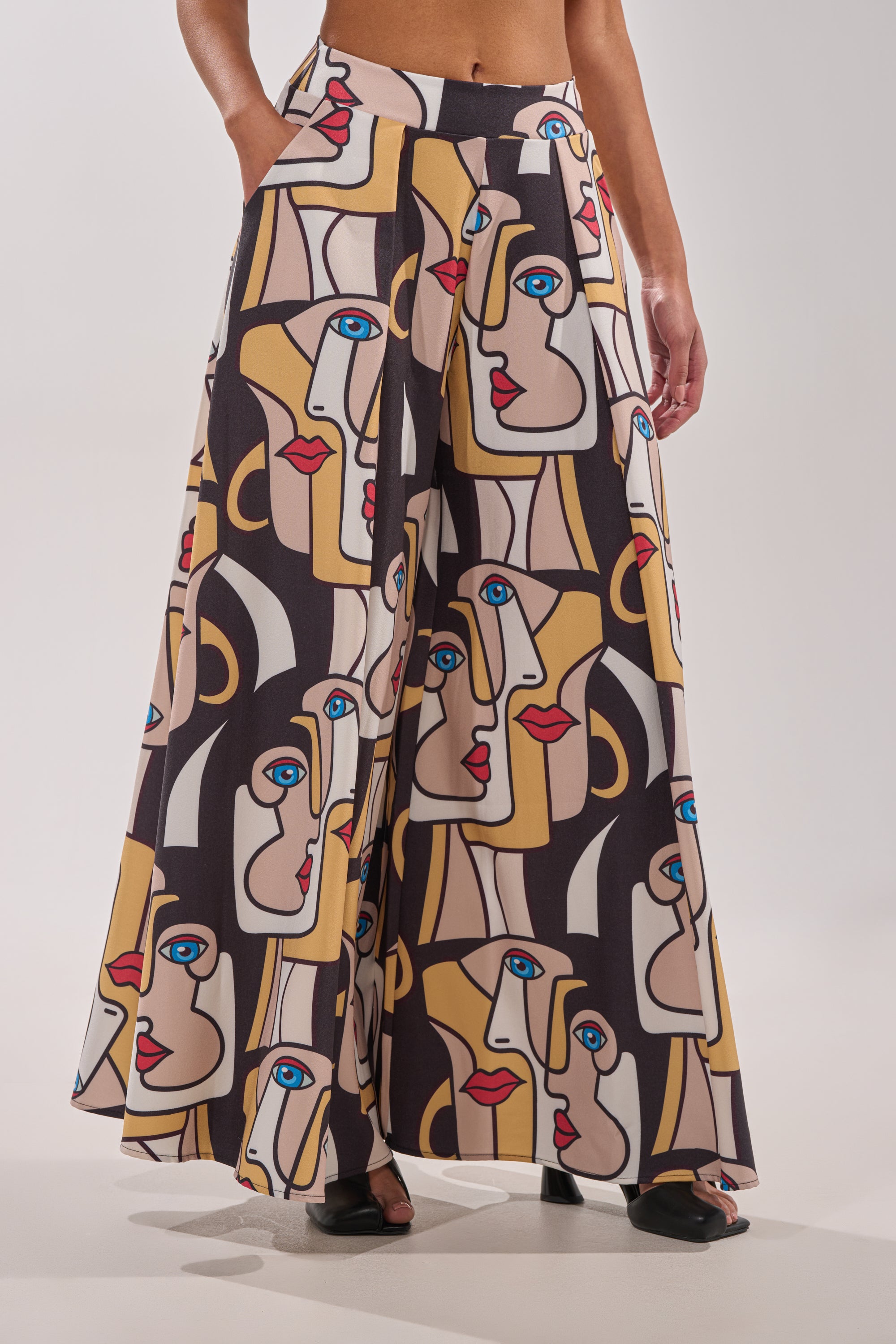 Wearing the FACES EVERYWHERE TROUSER, featuring wide legs and a bold abstract face print in beige, black, blue, red, and yellow; model poses with hands in pockets, showing only lower torso and legs. 0